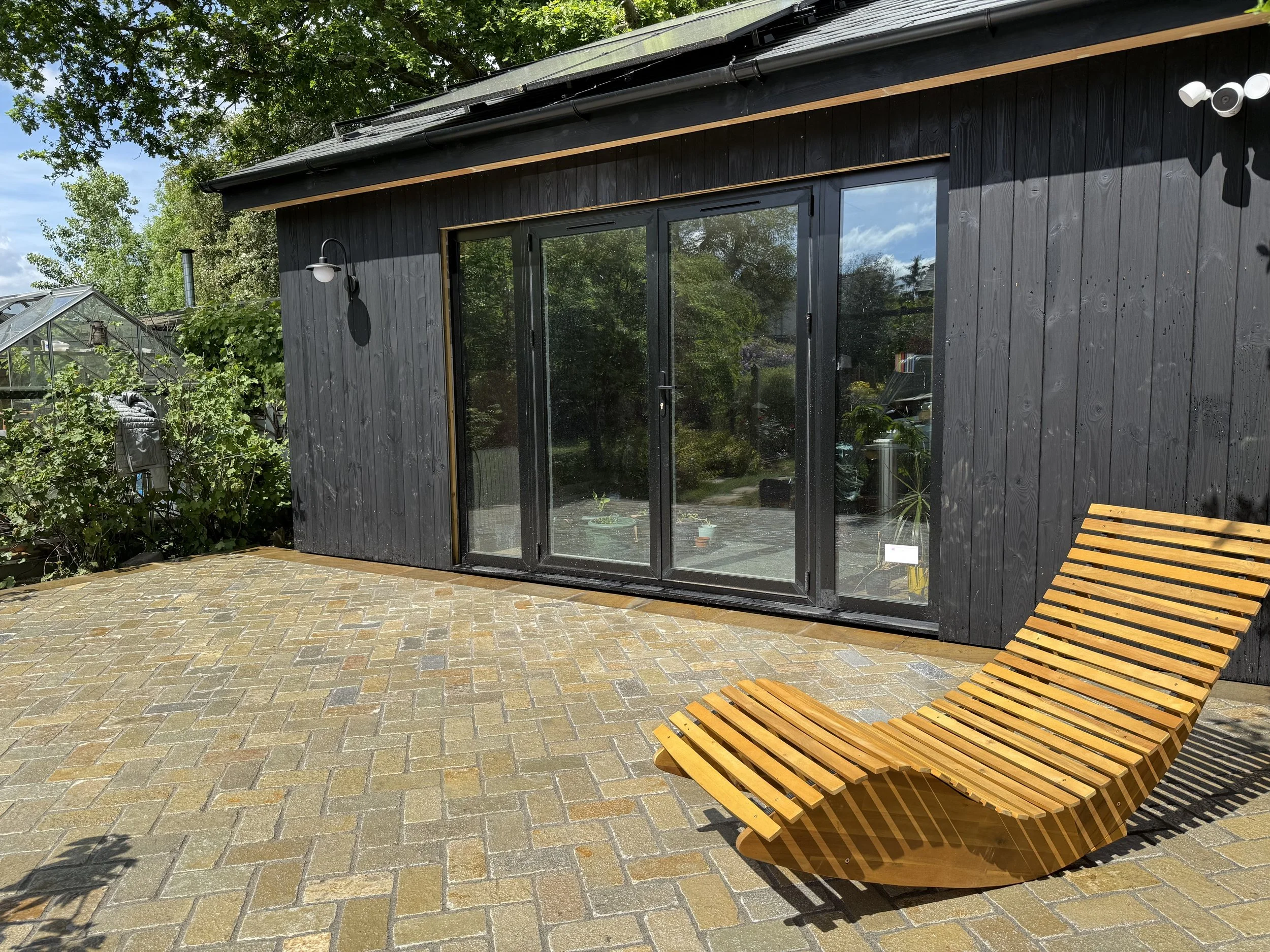 A black modern house with a sliding glass door, outdoor wooden lounge chair, and garden with green plants and a greenhouse under a leafy tree on a brick patio.