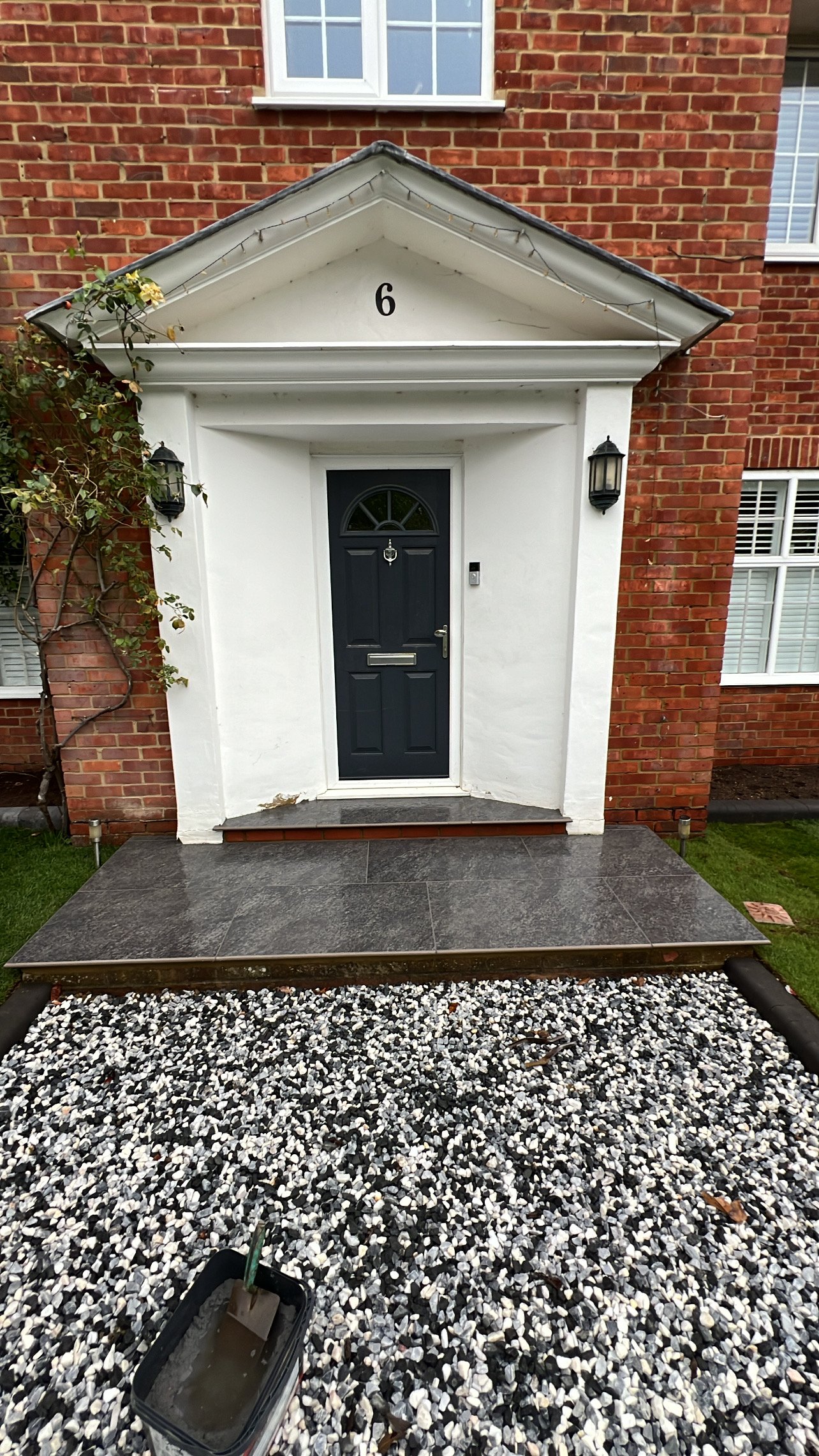 Front entrance of a brick house with a black door, white surrounding walls, and a tiled porch. There is a flowerpot with water and a paintbrush, indicating gardening or maintenance work, and white and black gravel on the ground.