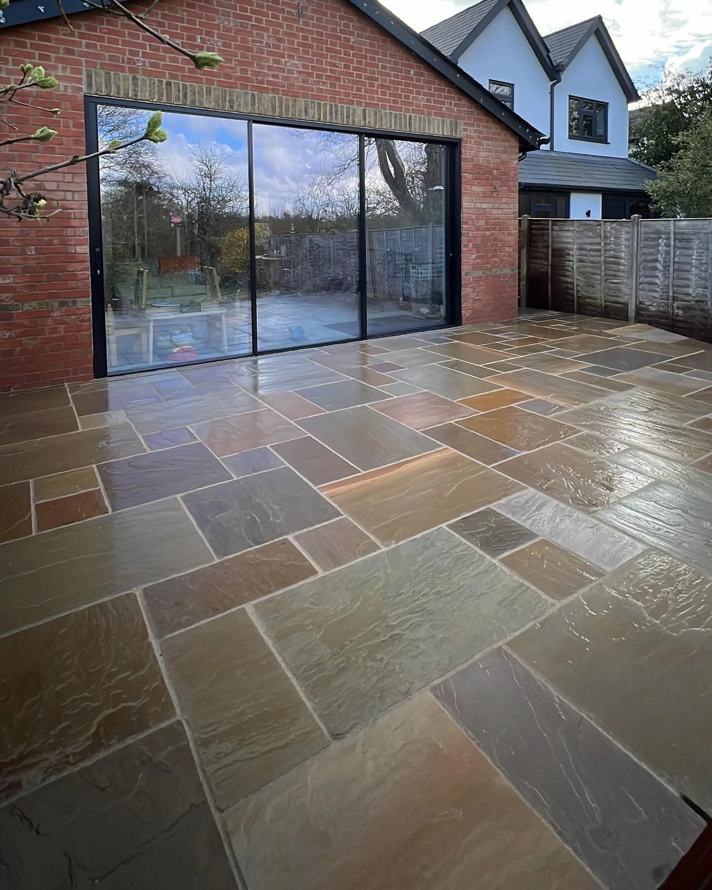 A courtyard with a wet stone tiled floor, a brick house with large glass sliding doors, a wooden fence, and a neighboring house with white and black exterior.
