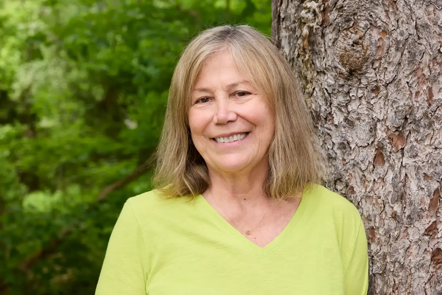 Dr. Anna Manatis, MD MPH, smiling outdoors, leaning against a tree trunk on Cape Cod.