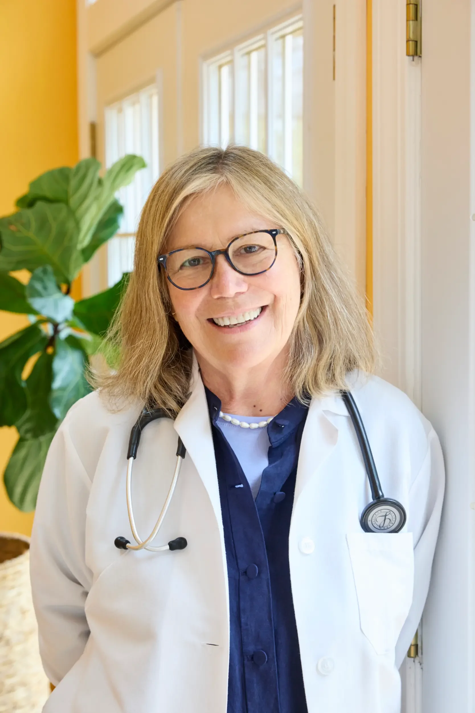 Dr. Anna Manatis, MD MPH, smiling in a white coat with a stethoscope around her neck, indoors near a bright window.