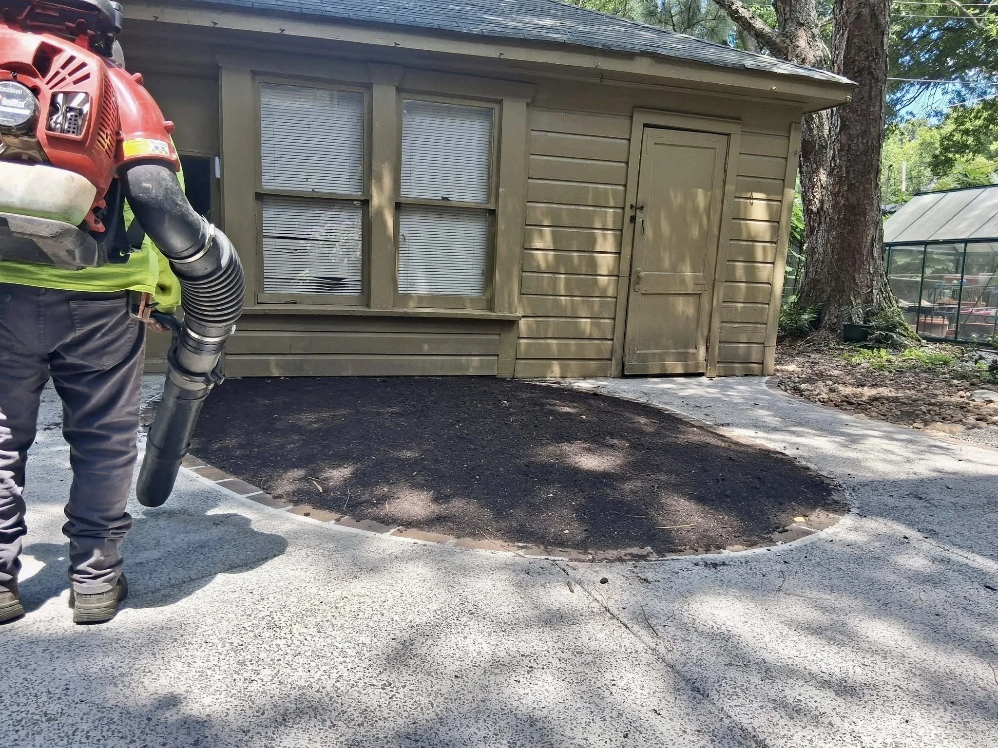 A person wearing a high-visibility vest and gray work pants, holding a leaf blower, standing on a freshly paved driveway in front of a small beige house with two windows and a door, surrounded by trees and a garden with a greenhouse.