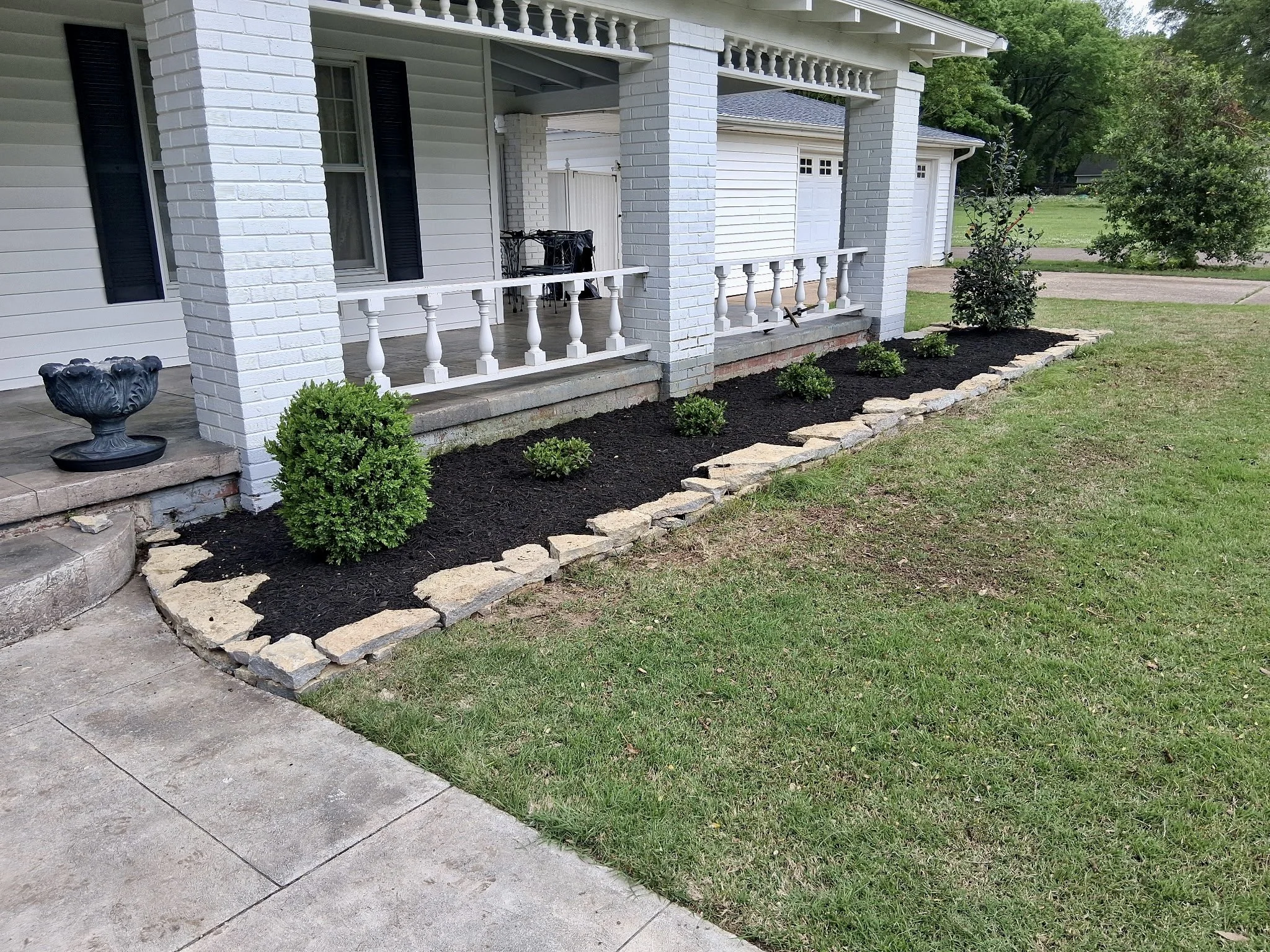 Front porch of a house with white brick pillars, black shutters, and a small garden bed with bushes and mulch. Visible part of the sidewalk and lawn.