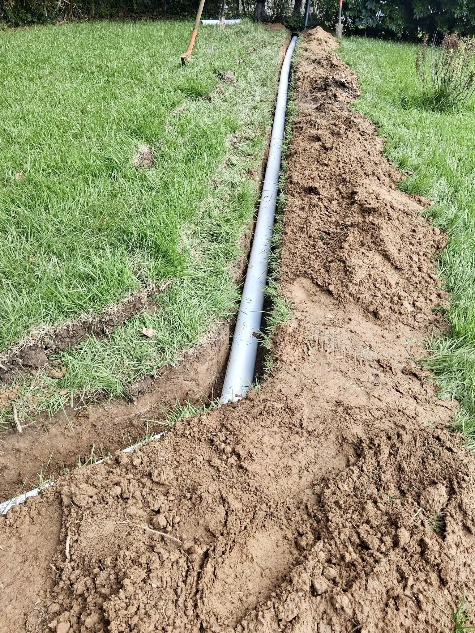 A trench in the ground with an underground pipe installed. The trench is surrounded by green grass and there is a pile of soil on the side.