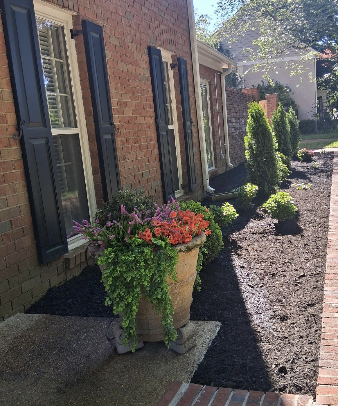 Brick house exterior with black shutters on windows, a potted plant with purple and orange flowers, and a freshly mulched garden bed with small bushes and trees.