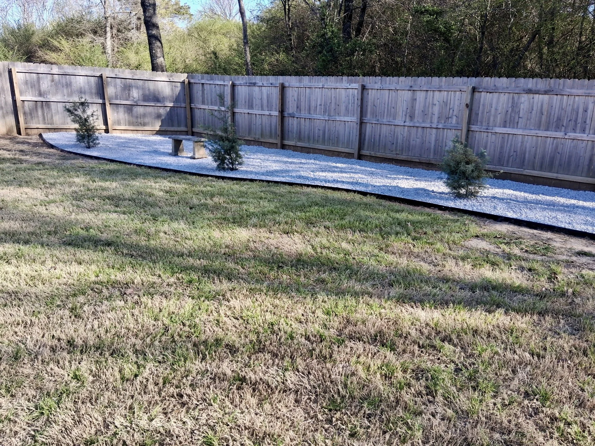 Backyard with a wooden privacy fence, small trees, a concrete bench, and a gravel border with grass in the foreground, surrounded by trees.