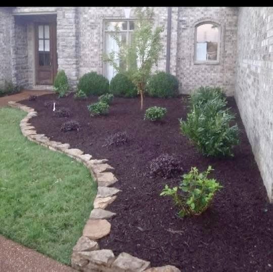 A landscaped garden bed with dark mulch, green bushes, and a small tree, bordered by stones, adjacent to a brick house with windows and a door.