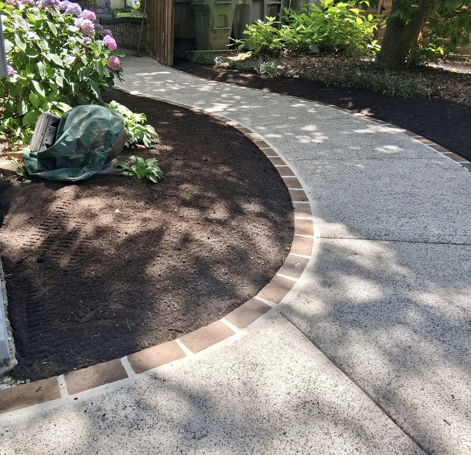 A garden pathway with newly laid brick border around a curved concrete path. There are plants and bushes on either side of the path, with some bushes flowering with pink blooms. A covered gardening tool or bag is visible on the soil next to the plants on the left.