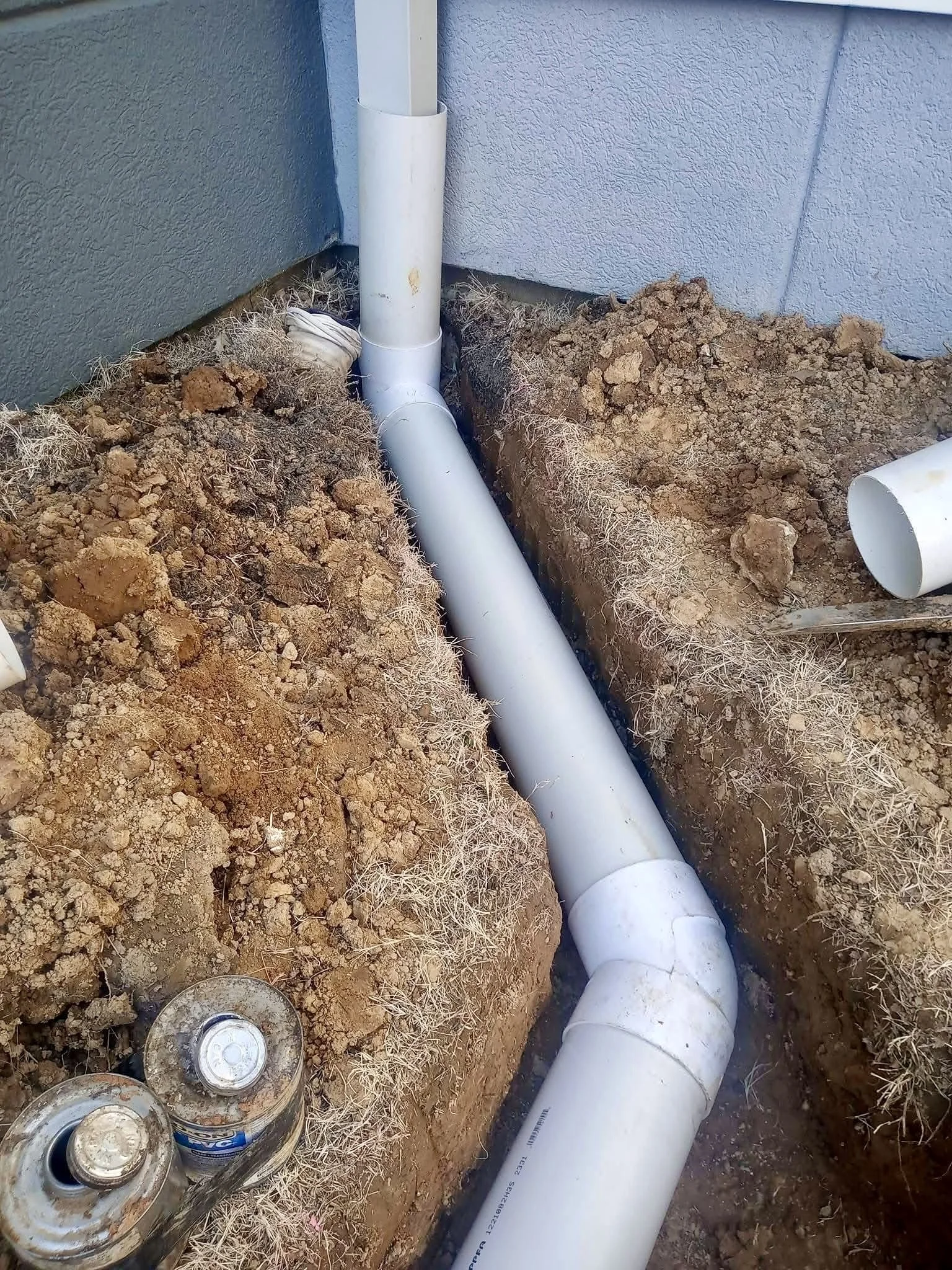 Pipes installed underground near the foundation of a building, with dirt and construction materials around.