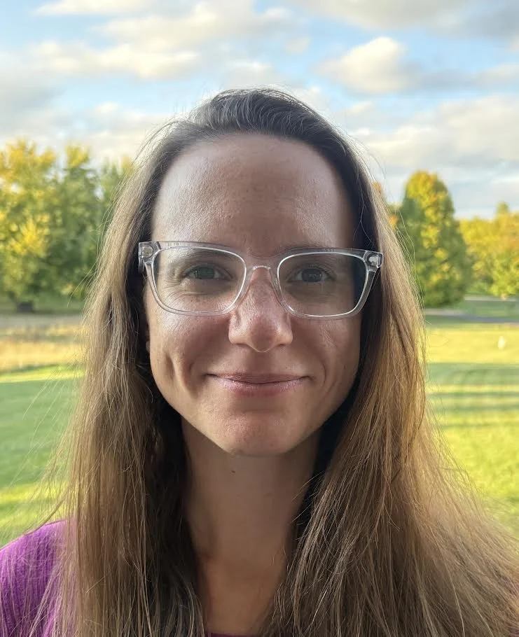 Close-up of a woman with long brown hair wearing glasses, smiling outdoors with trees and a blue sky in the background.
