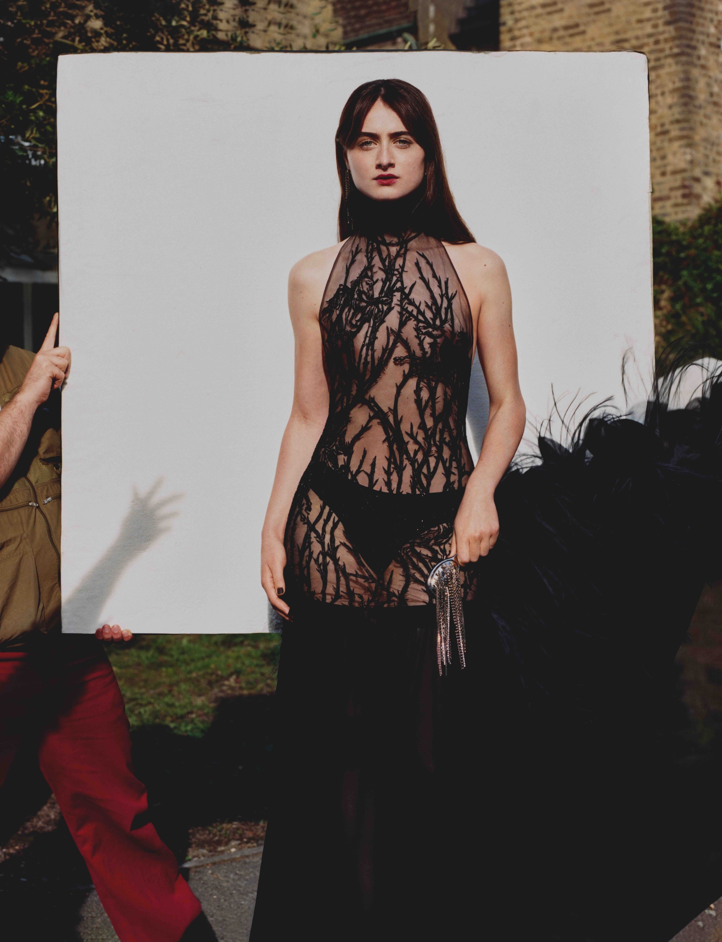 A woman with long, dark hair wearing a sheer, black dress with branch-like patterns stands in front of a white panel at an outdoor event. A person in red pants is holding the panel, and there are other people and buildings in the background.