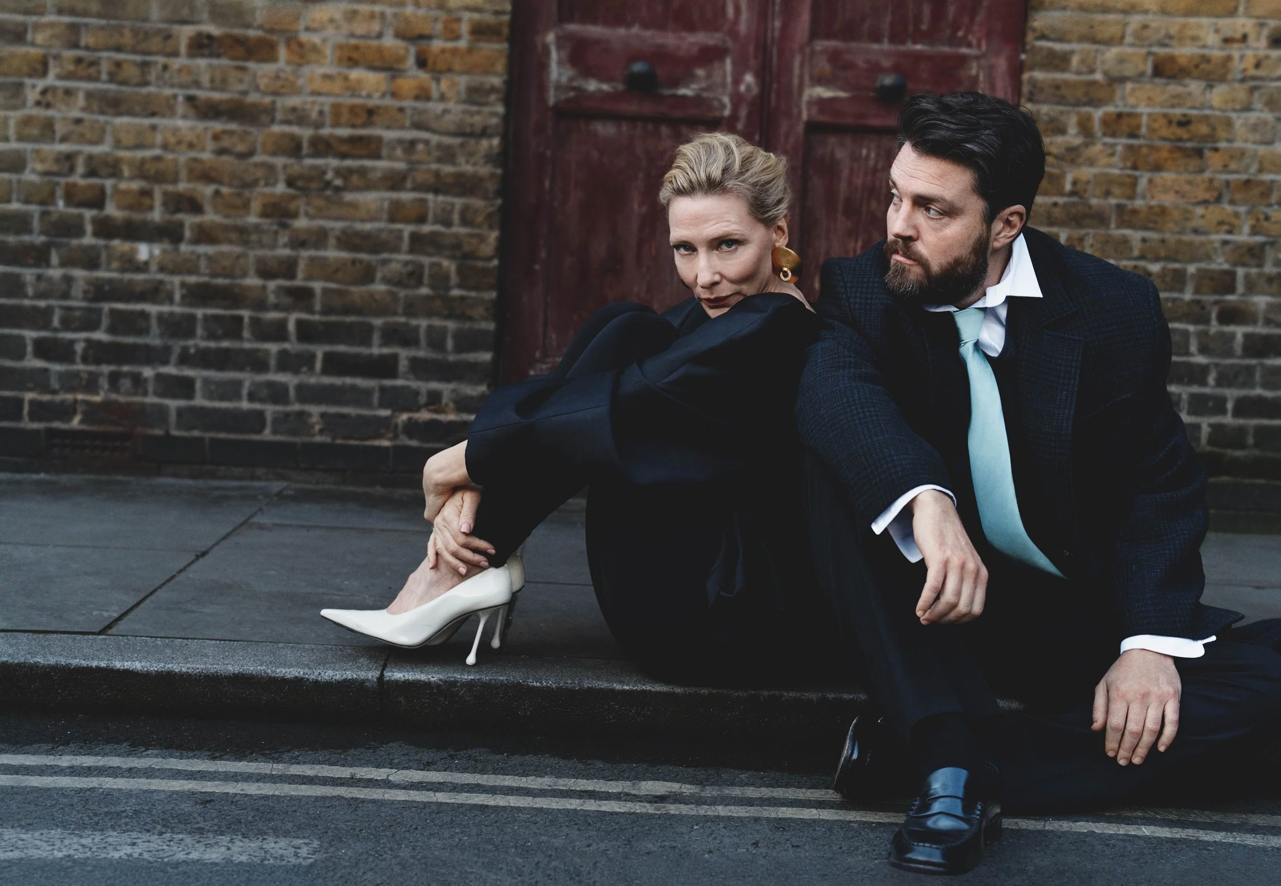 A woman with blonde hair and a man with dark hair and a beard sit on a sidewalk in front of a brick wall with a red door. The woman is wearing white high heels, earrings, and a dark blazer and is leaning on the man's shoulder. The man is wearing a dark suit with a white shirt and a light blue tie, and is looking to his right.