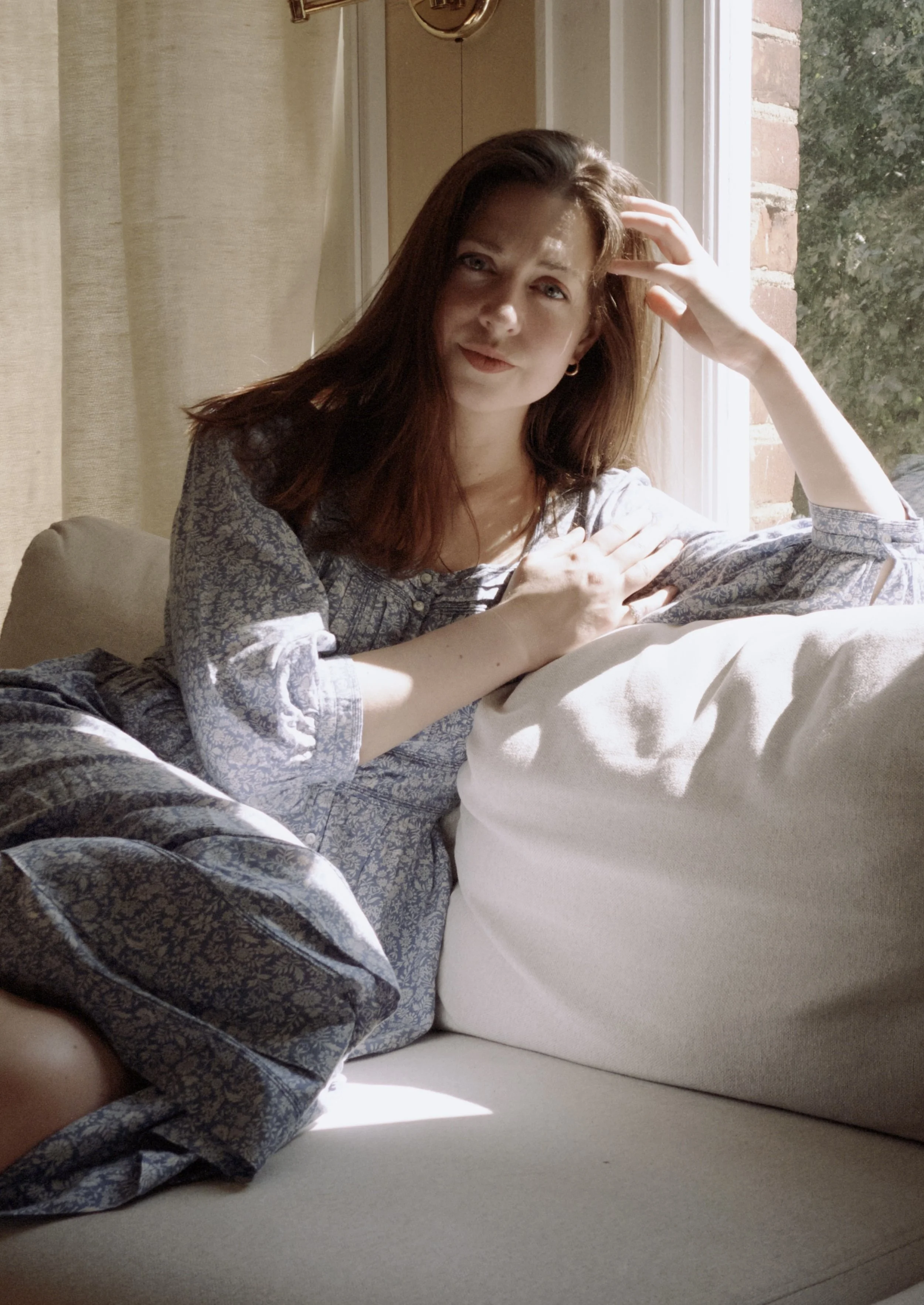 A young woman with red hair and light skin sitting on a beige sofa near a window, with sunlight shining on her. She is leaning against a pillow, resting her hand on her head, and looking directly at the camera.