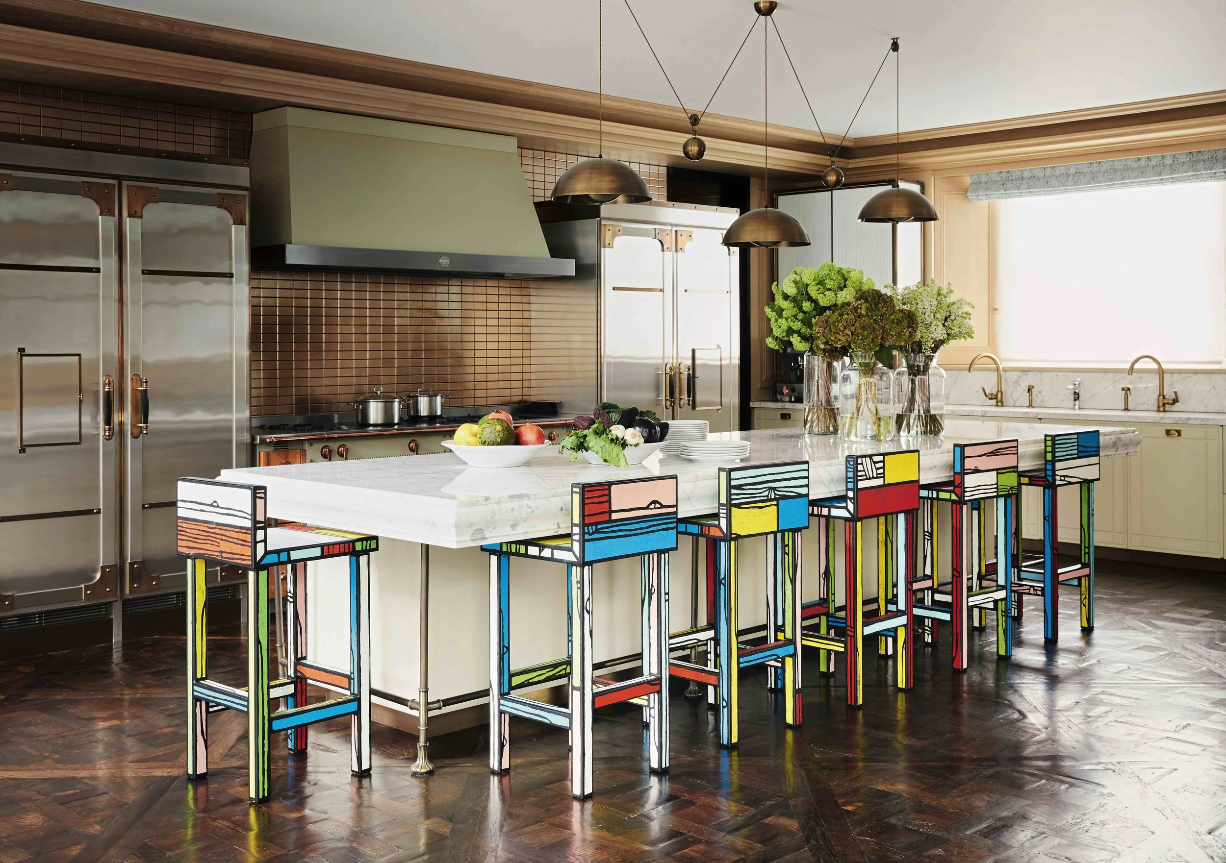 A modern kitchen with a white marble island counter, decorated with colorful geometric-patterned stools. The kitchen features stainless steel appliances, a brown tile backsplash, green and beige cabinetry, and a large window with a Roman shade. A bouquet of green and brown flowers is on the countertop.
