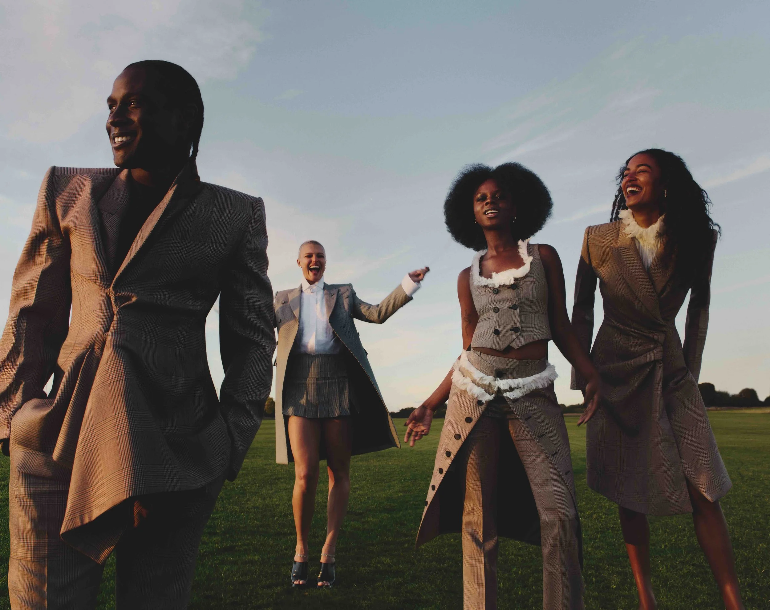 A diverse group of four stylishly dressed people walking outdoors on a grass field during sunset, smiling and enjoying each other's company.