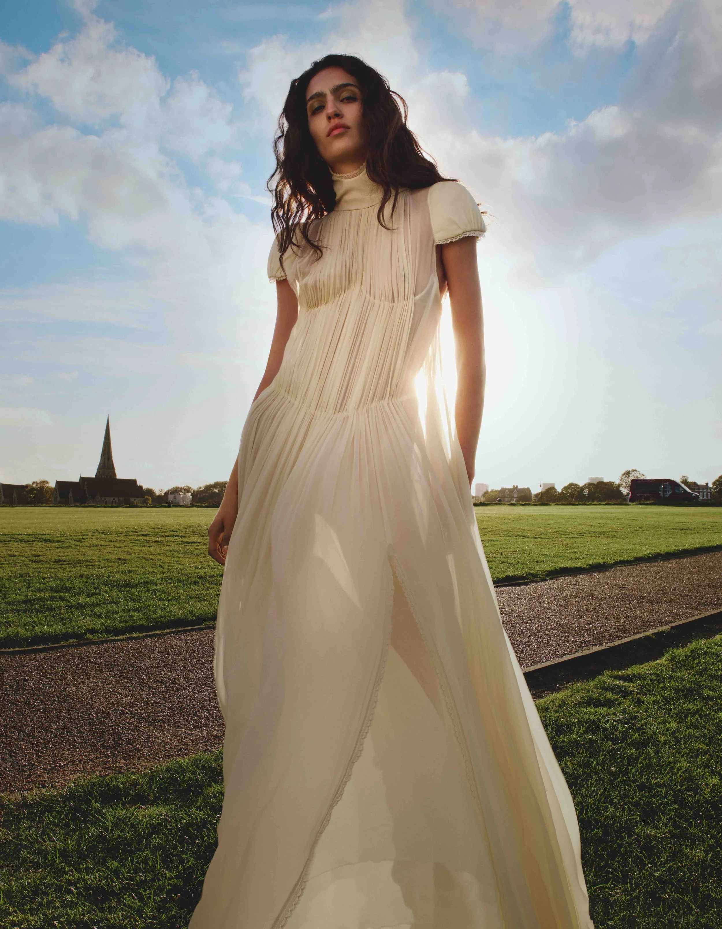 A woman with long wavy dark hair wearing a beige dress stands outdoors in a grassy field with a church steeple and a row of houses in the background, under a partly cloudy sky.
