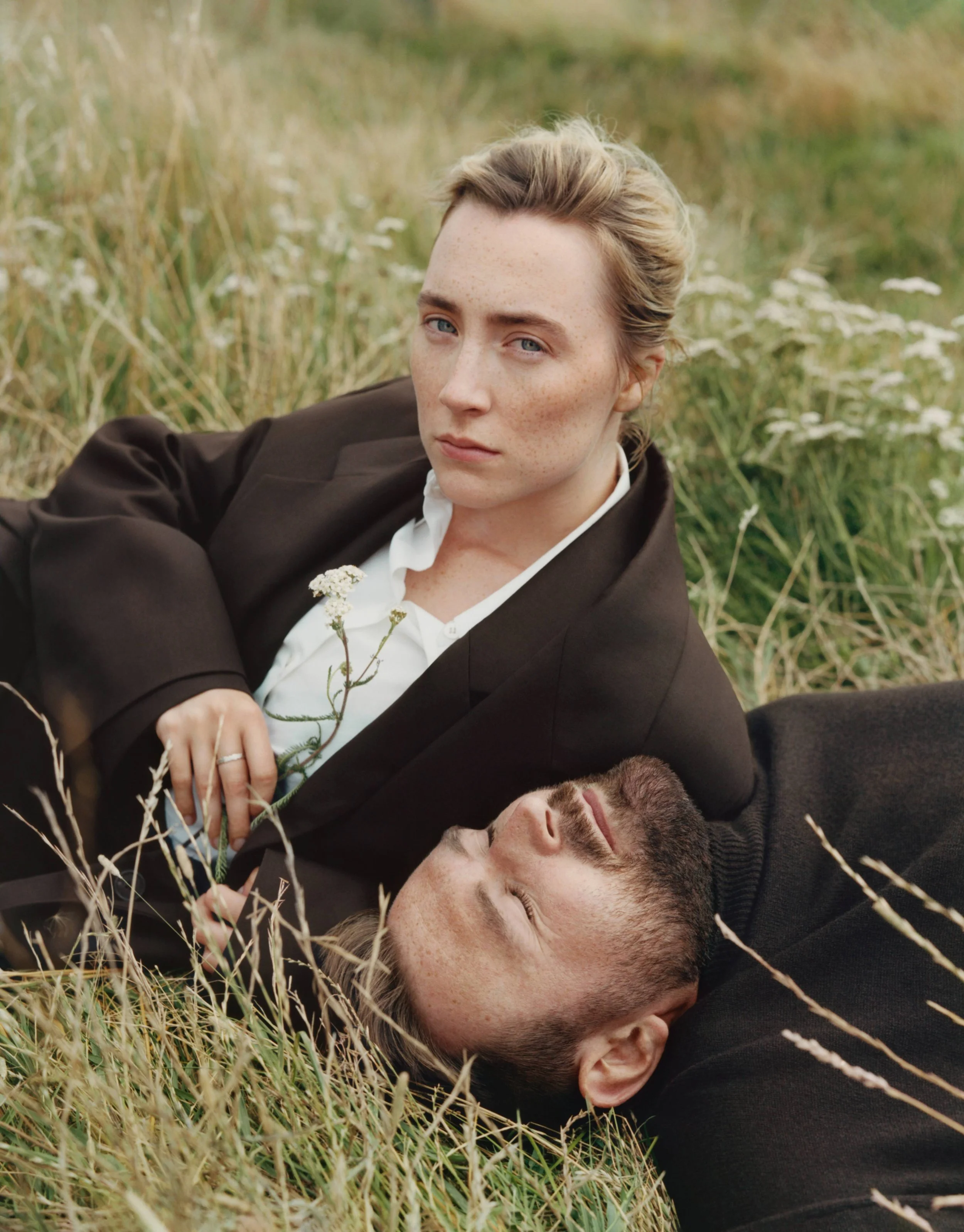 A woman with blonde hair and freckles lying in tall grass, wearing a black blazer and white shirt, with a man lying beside her, both appearing relaxed in a natural outdoor setting.
