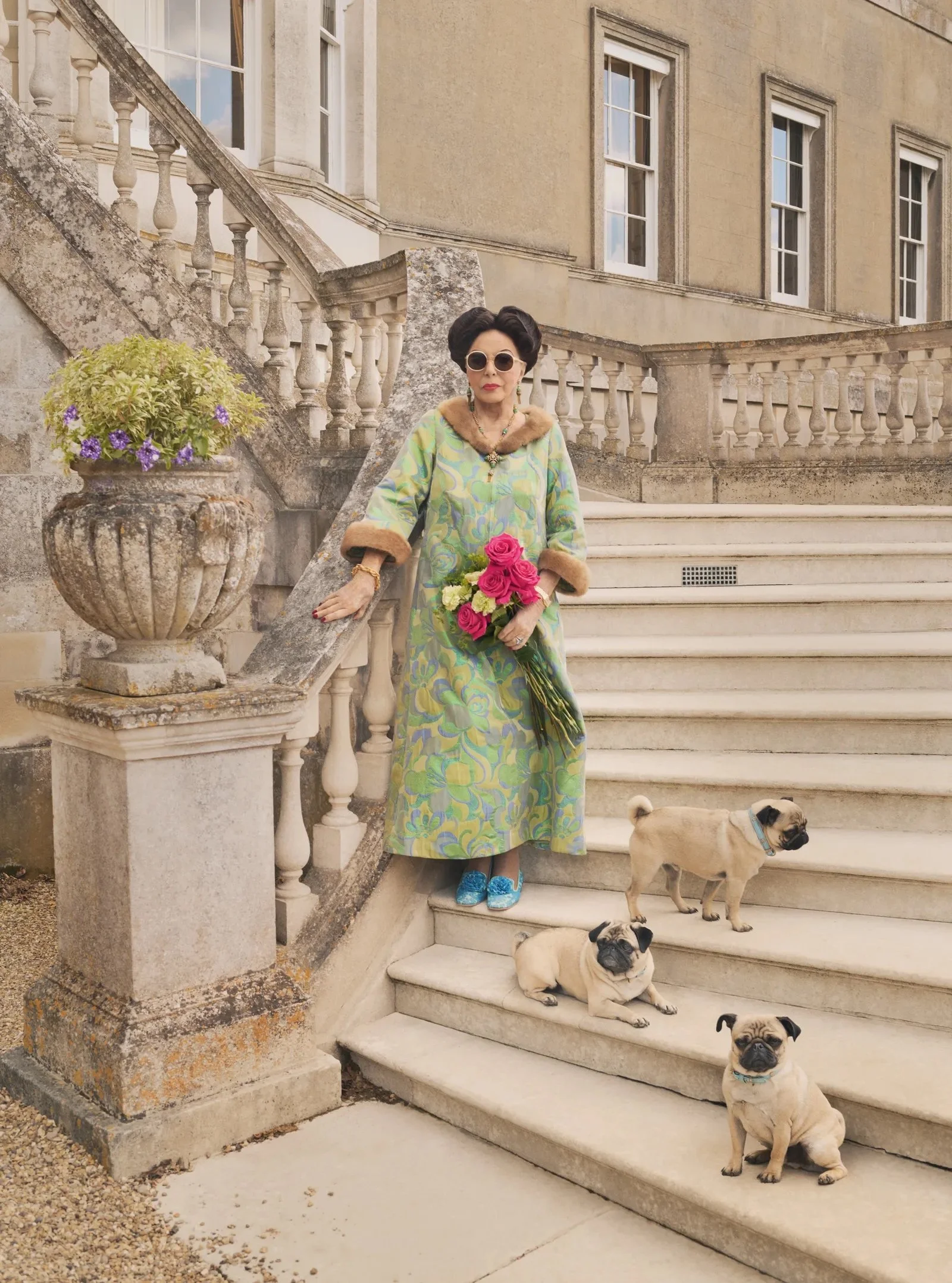 A woman in vintage clothing and sunglasses holding a bouquet of pink roses standing on stone stairs with four pugs sitting nearby and a historic building in the background.