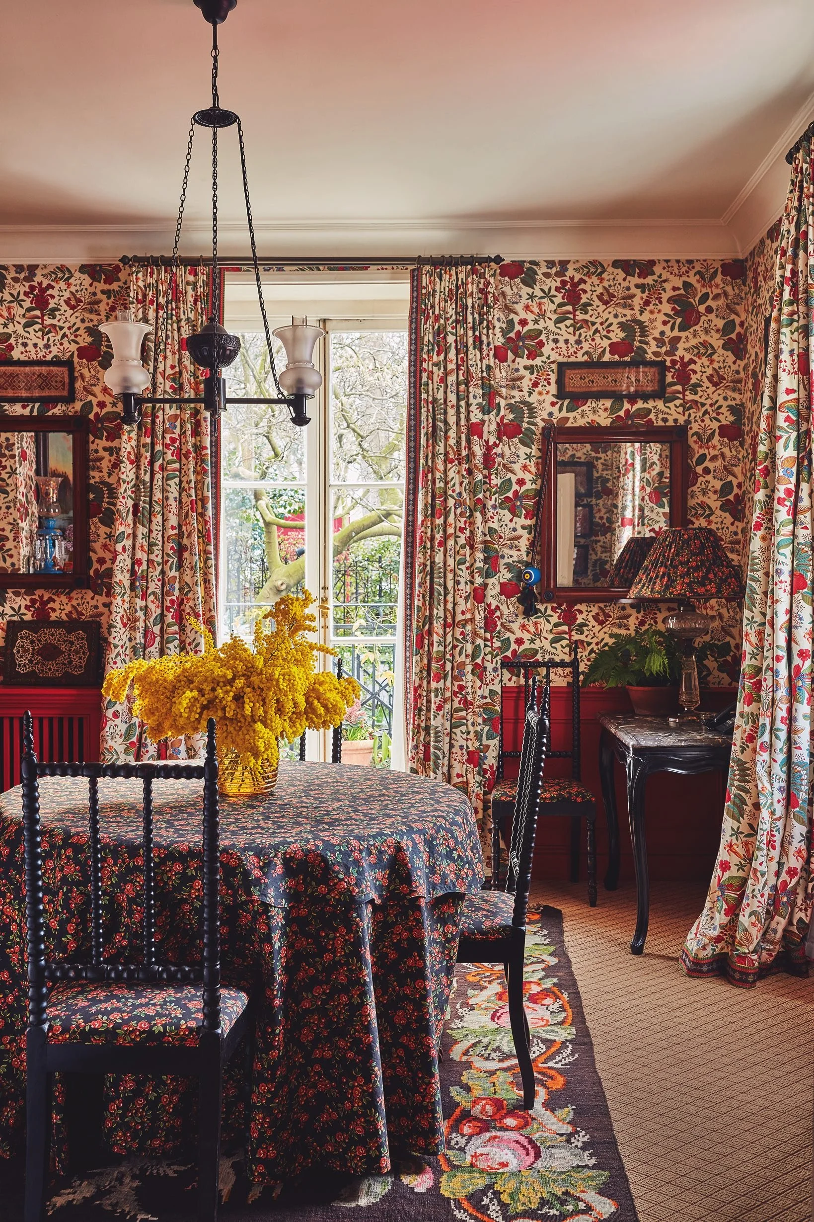 A cozy dining room with floral wallpaper and patterned curtains. A table with a dark floral tablecloth is centered, decorated with a large yellow floral centerpiece. Surrounding chairs have matching floral upholstery. A window with open curtains shows a view of trees outside. The room features a black side table with decorative items and a table lamp with a floral shade. A vintage chandelier hangs from the ceiling.