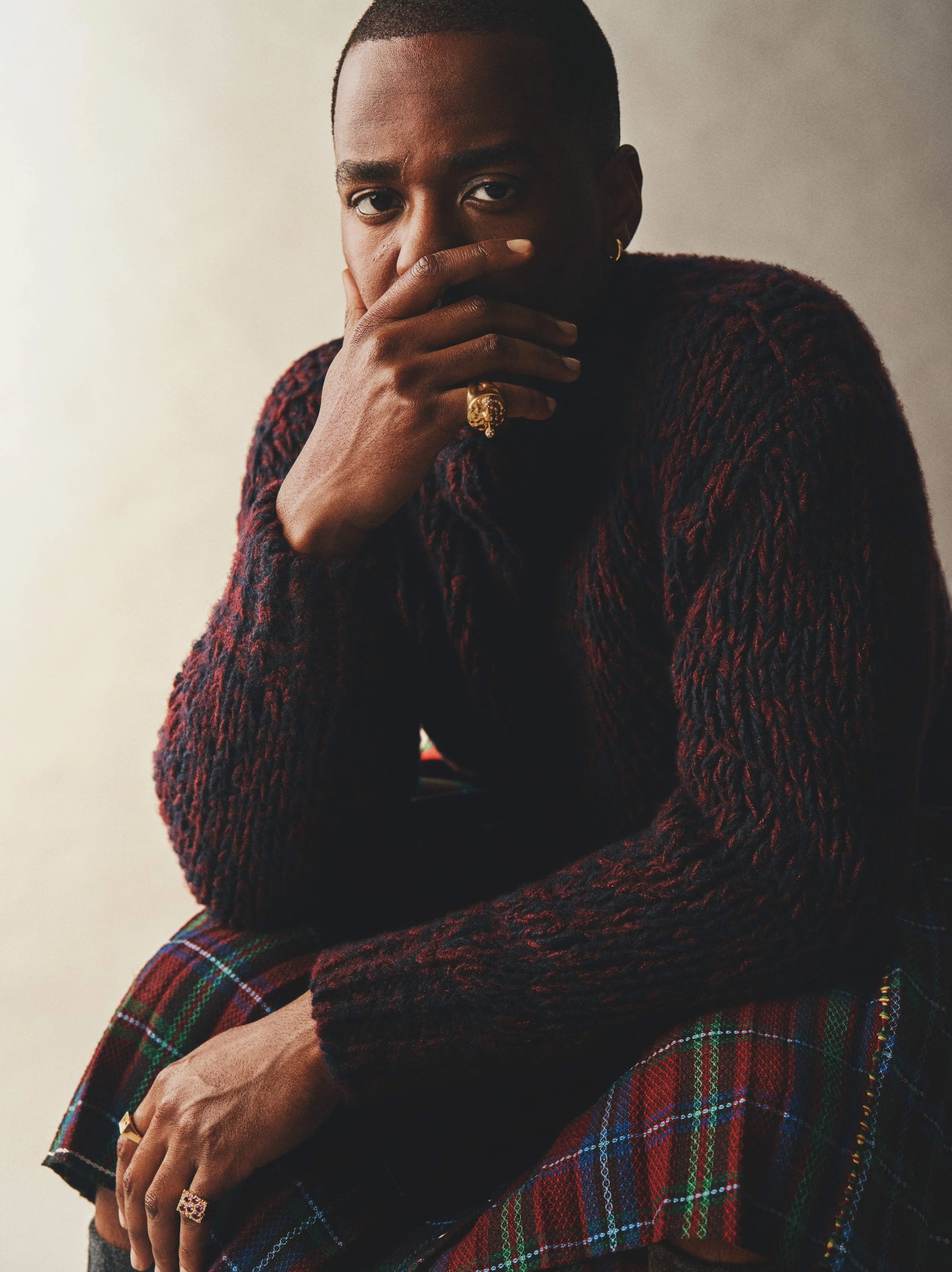 Portrait of a person with short hair wearing a dark red textured sweater and plaid skirt, with gold jewelry including rings and a hoop earring, sitting with one arm crossed and hand touching face, against a plain background.