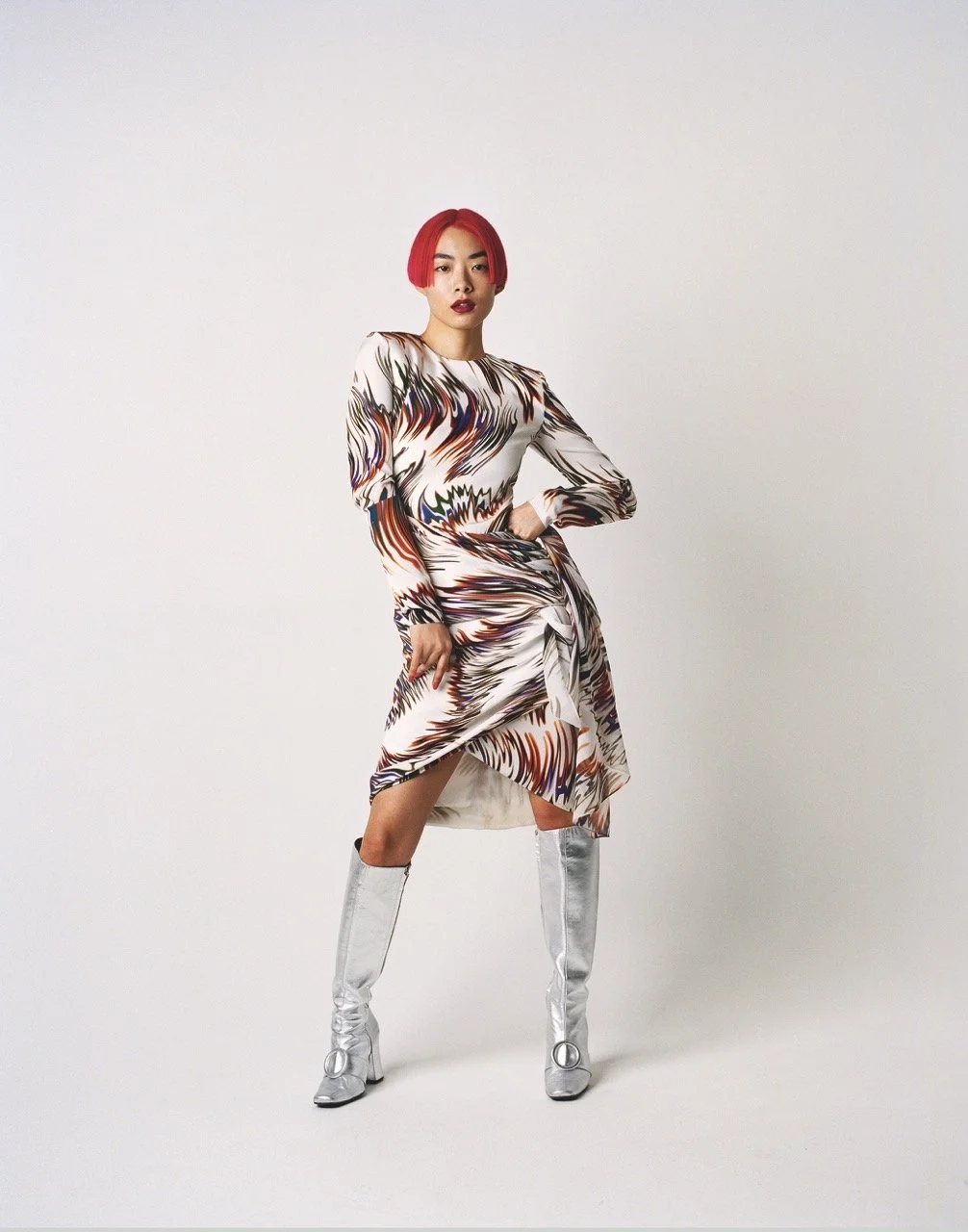 A woman with bright red hair styled in a bob, wearing a long-sleeved, multicolored, abstract patterned dress that is knee-length with a slit, paired with knee-high silver boots, standing against a plain, light background.