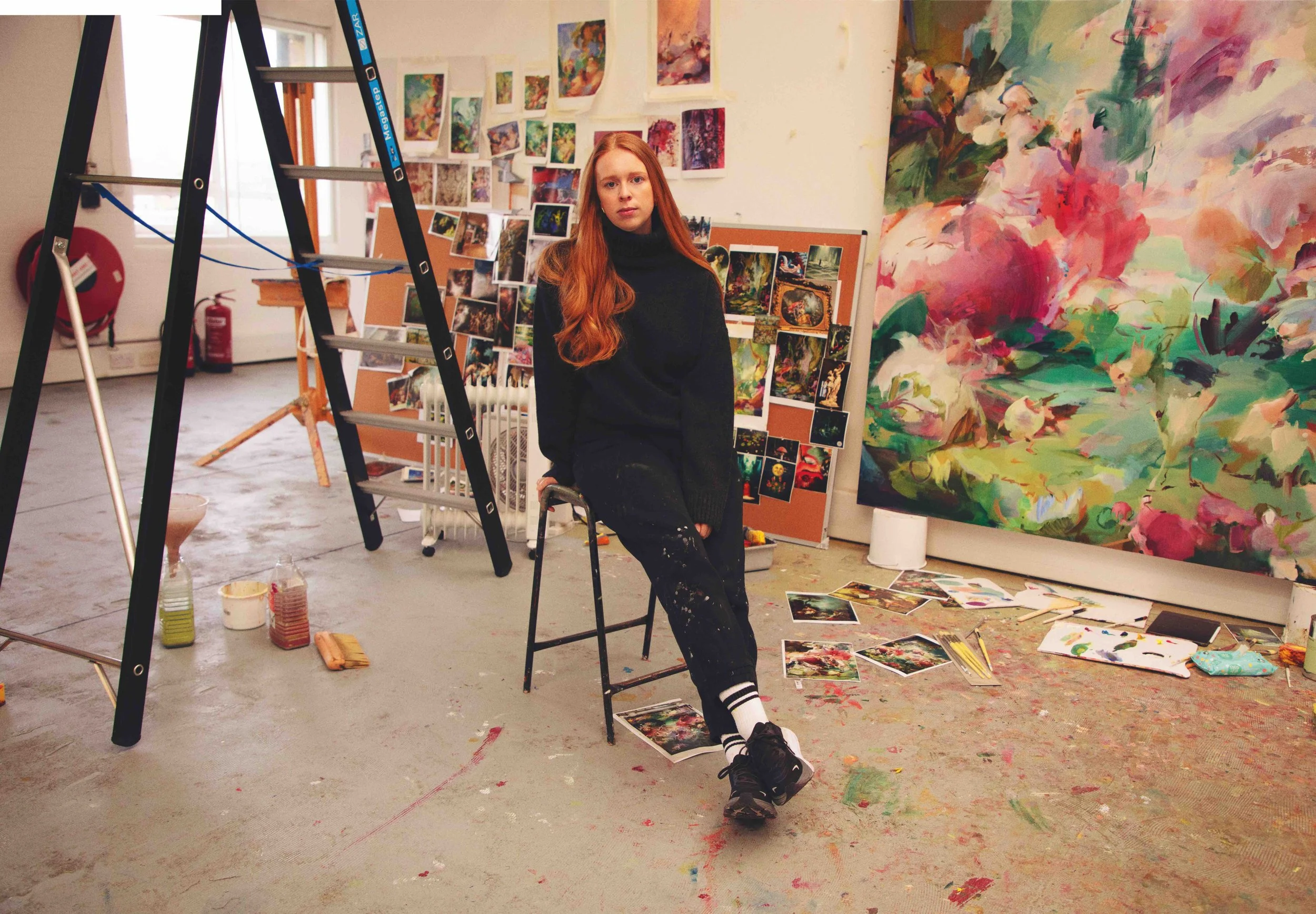 A young woman with long red hair sitting on a black chair in an art studio surrounded by colorful paintings, photographs, and art supplies, with a large abstract floral painting in the background.