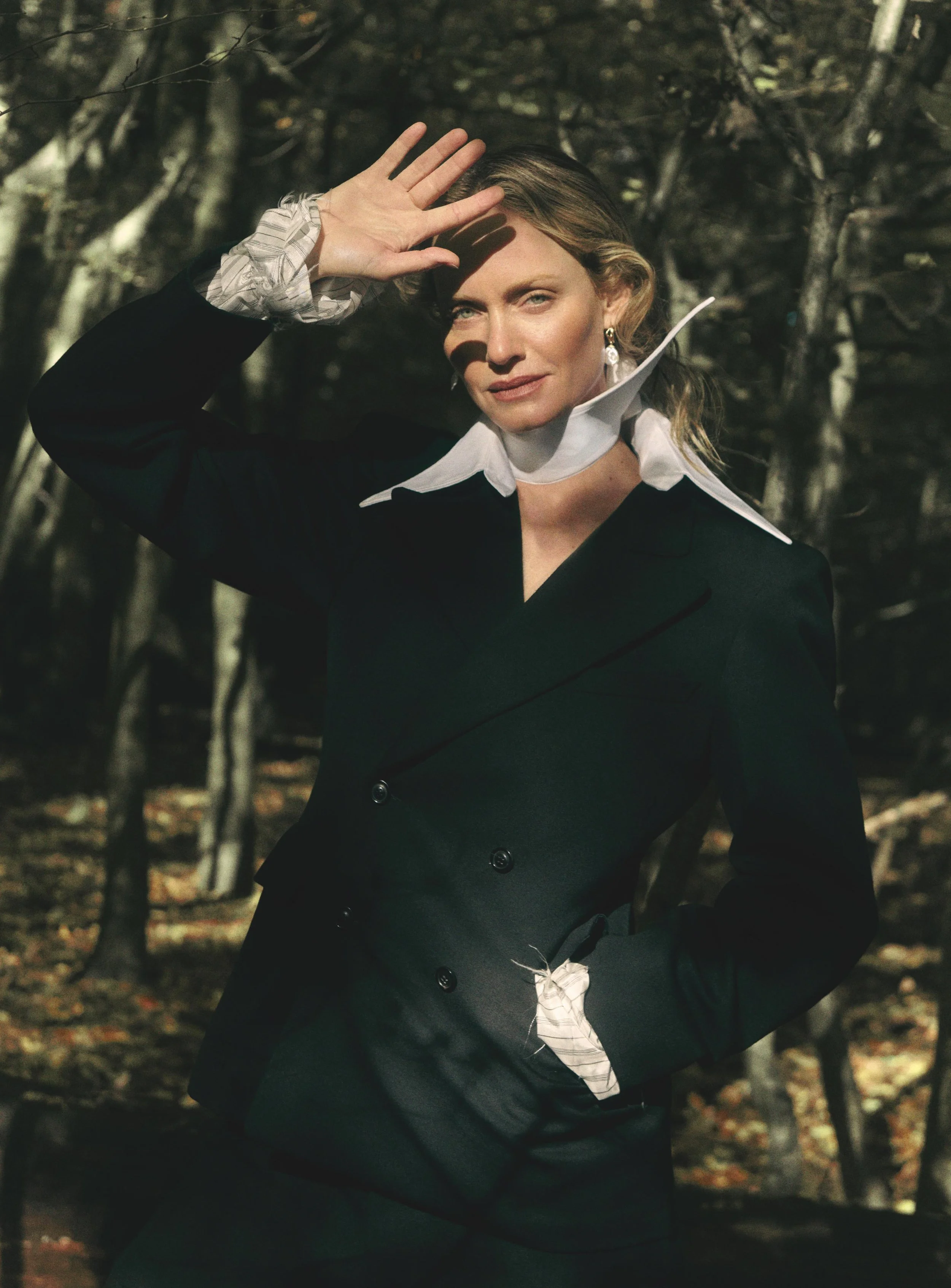 A woman in a black suit and jewelry, with a white shirt and ruffled cuffs, blocking sunlight with her hand, in a wooded area with bare trees.