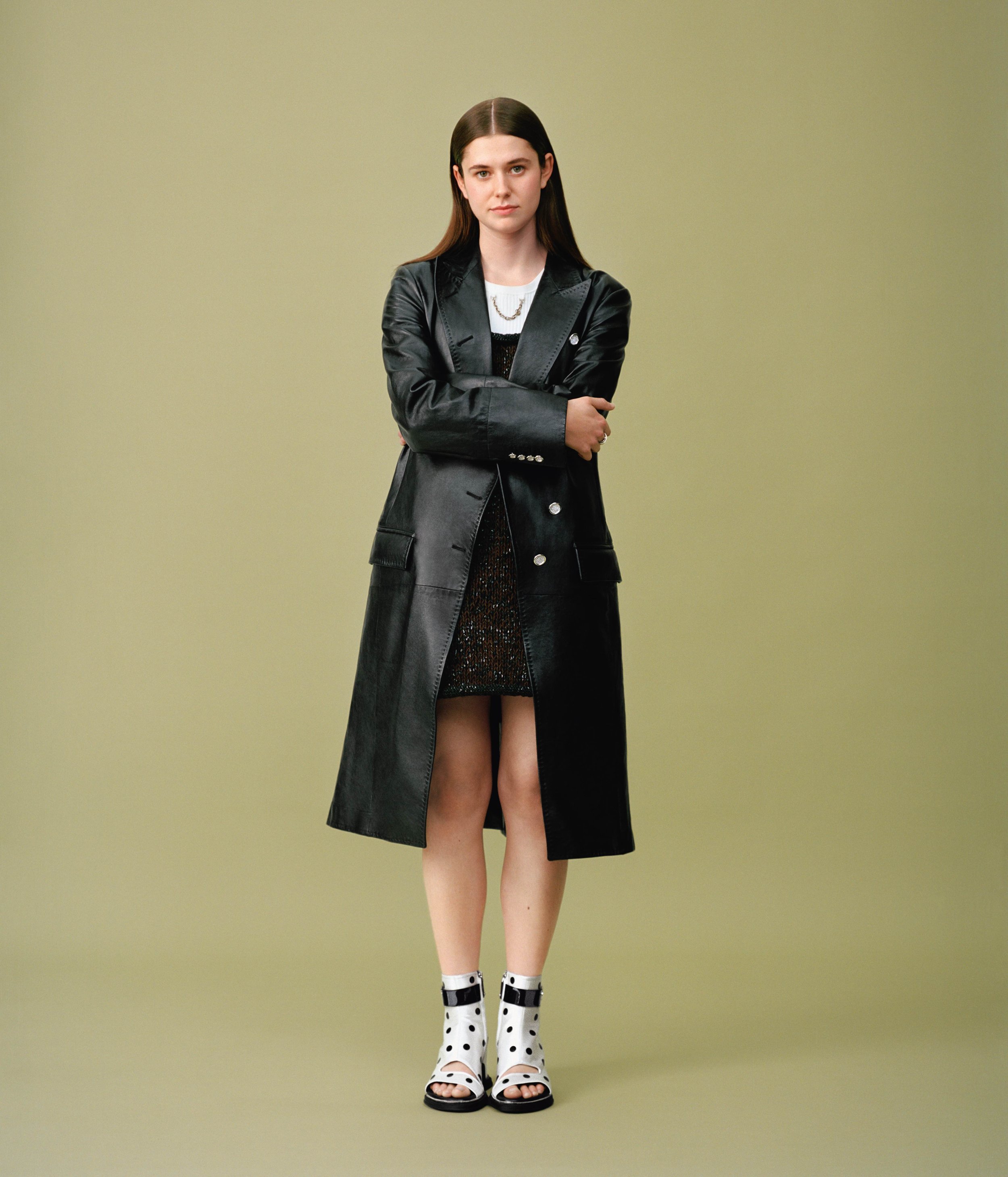 A young woman with long, dark hair wearing a black leather coat over a black dress, patterned socks, and polka dot high-heeled sandals poses against a plain, light green background.