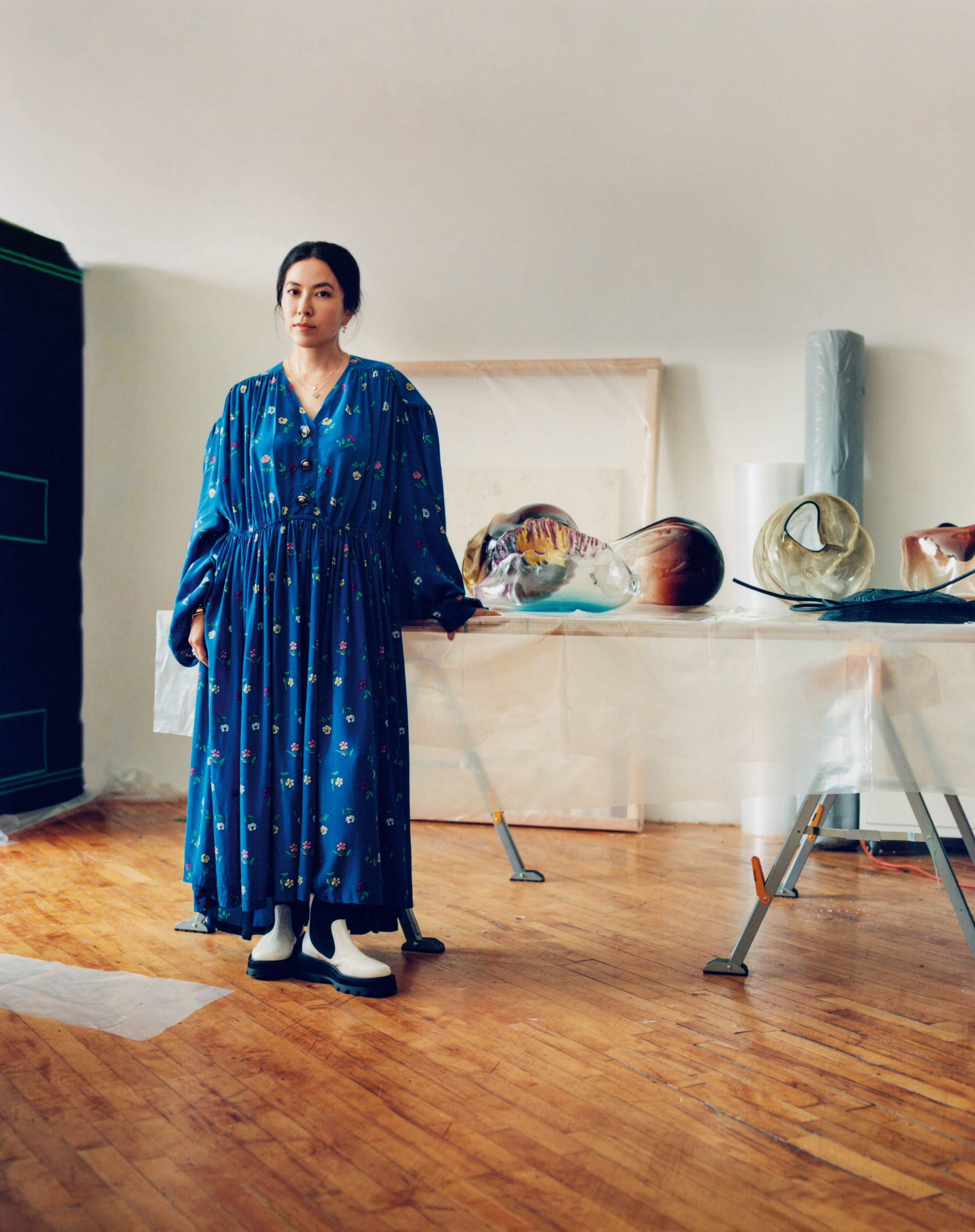 Woman standing in an art studio with glass sculptures on a table behind her, wearing a long blue floral dress and white boots.