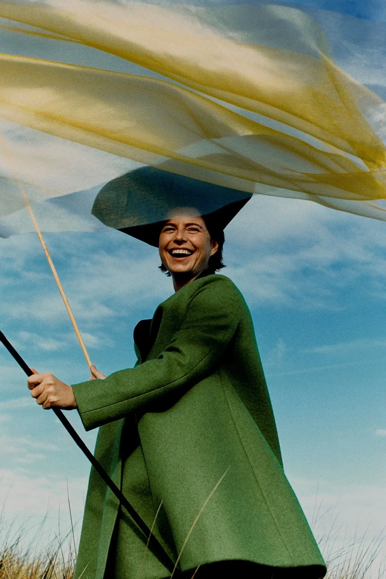 A woman smiling outdoors under a large yellow and white umbrella, wearing a green coat.