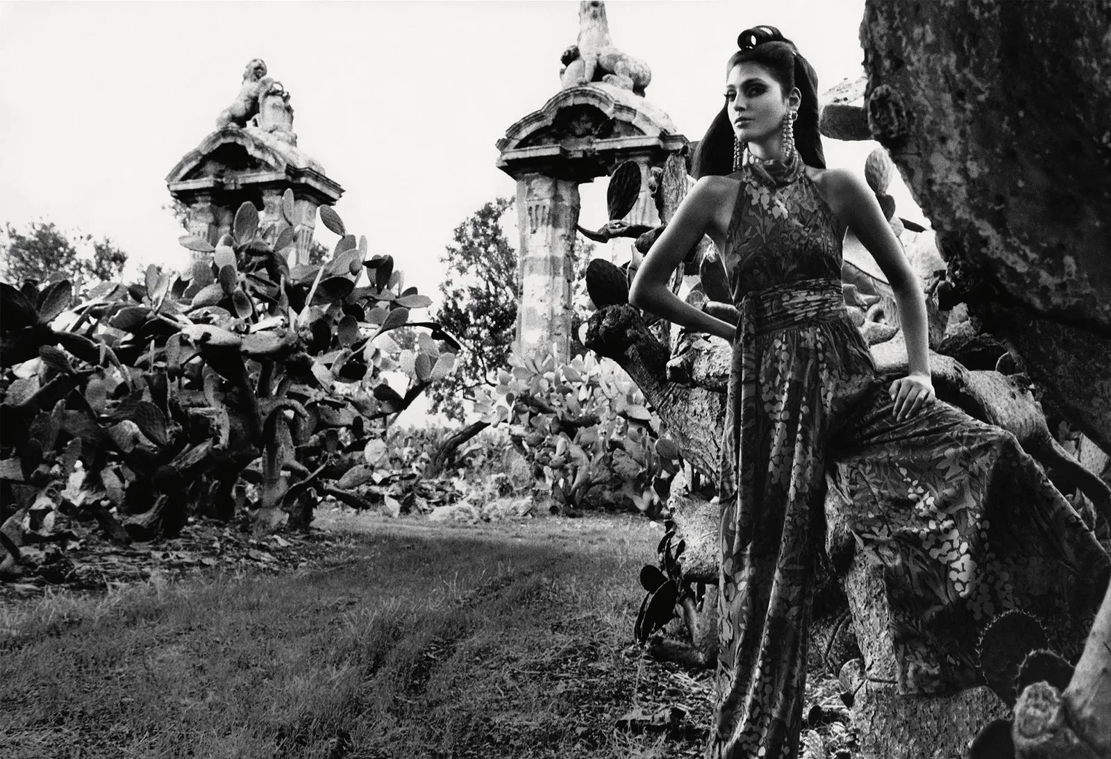 A woman in a patterned flowing dress with jewelry poses outdoors amongst rocks, trees, and old stone structures in black and white.