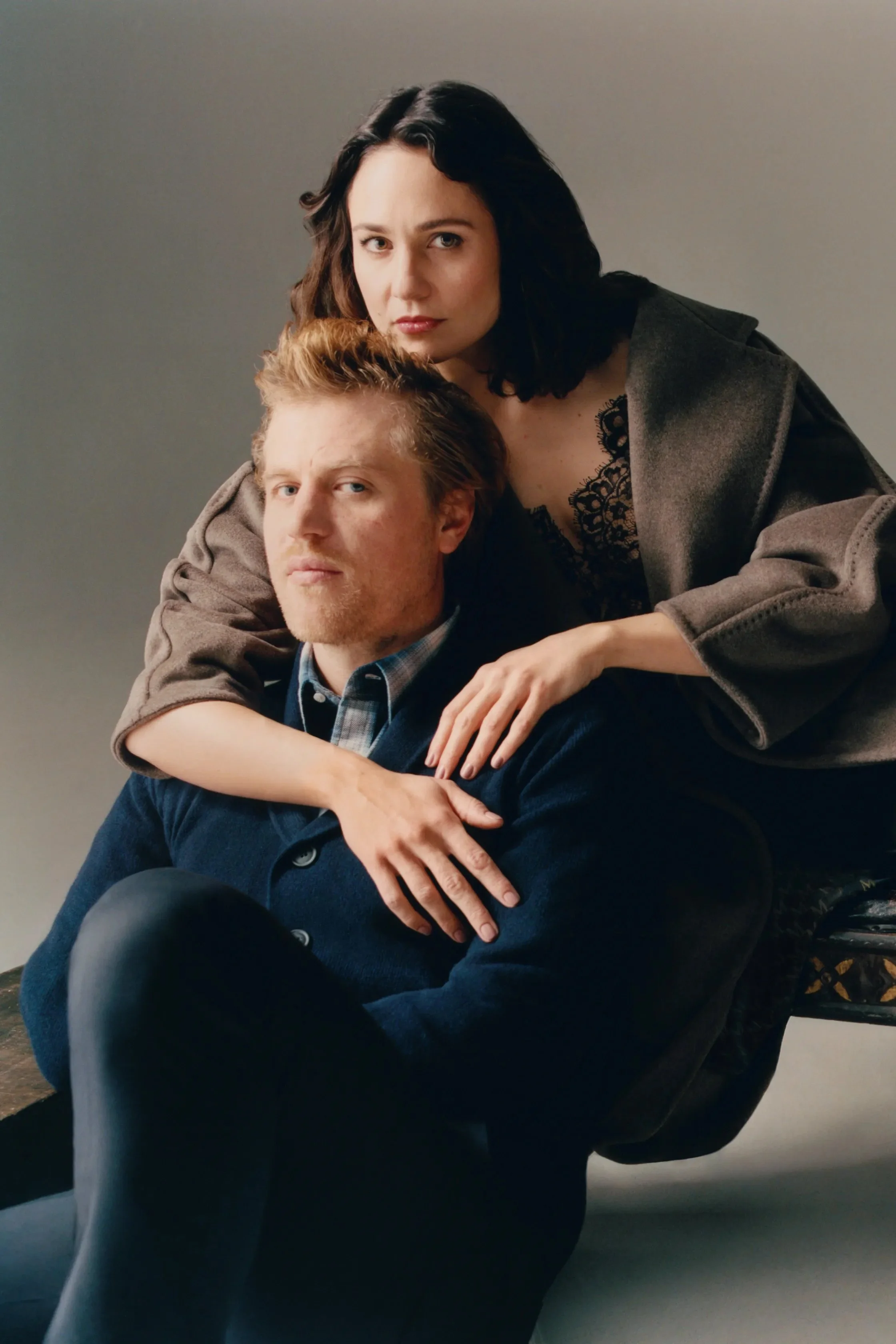 A woman with dark, wavy hair and light skin leaning over a man with light skin, reddish hair, and a beard, who is sitting on a black chair and wearing a dark blue sweater. The woman is wearing a dark jacket and lace top. They are against a plain, neutral-colored background.