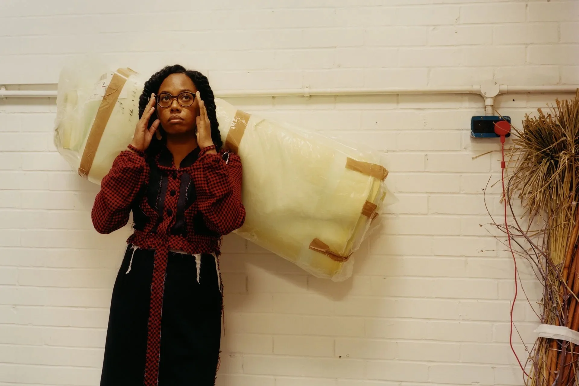 A woman with dark hair in braids, wearing glasses, a red and black checkered shirt, and a long black skirt, stands in front of a white brick wall. She is holding her head with both hands and has a serious expression. Behind her is a large yellow infl