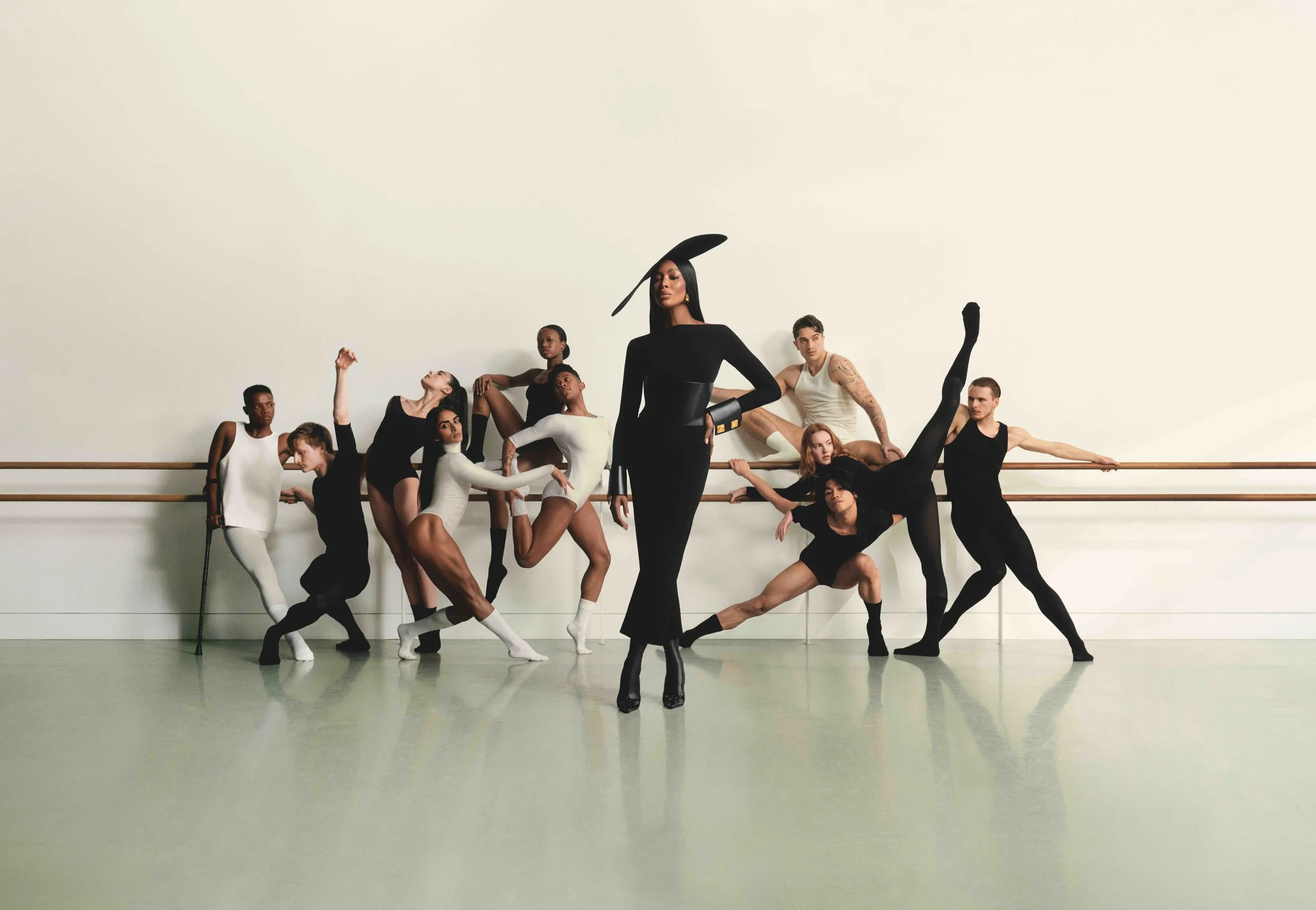 A diverse group of ballet dancers and a fashionably dressed woman posing in a dance studio with a ballet barre, some in classical ballet attire and others in black dancewear, with one dancer holding a cane and all striking various ballet poses.