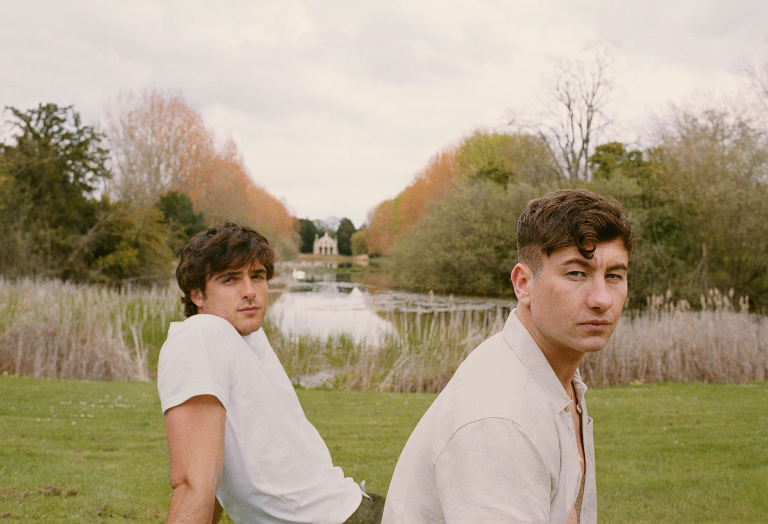 Two young men standing outdoors near a pond with trees in the background. One has dark wavy hair and the other has short curly hair. Both are wearing light-colored shirts and looking at the camera.