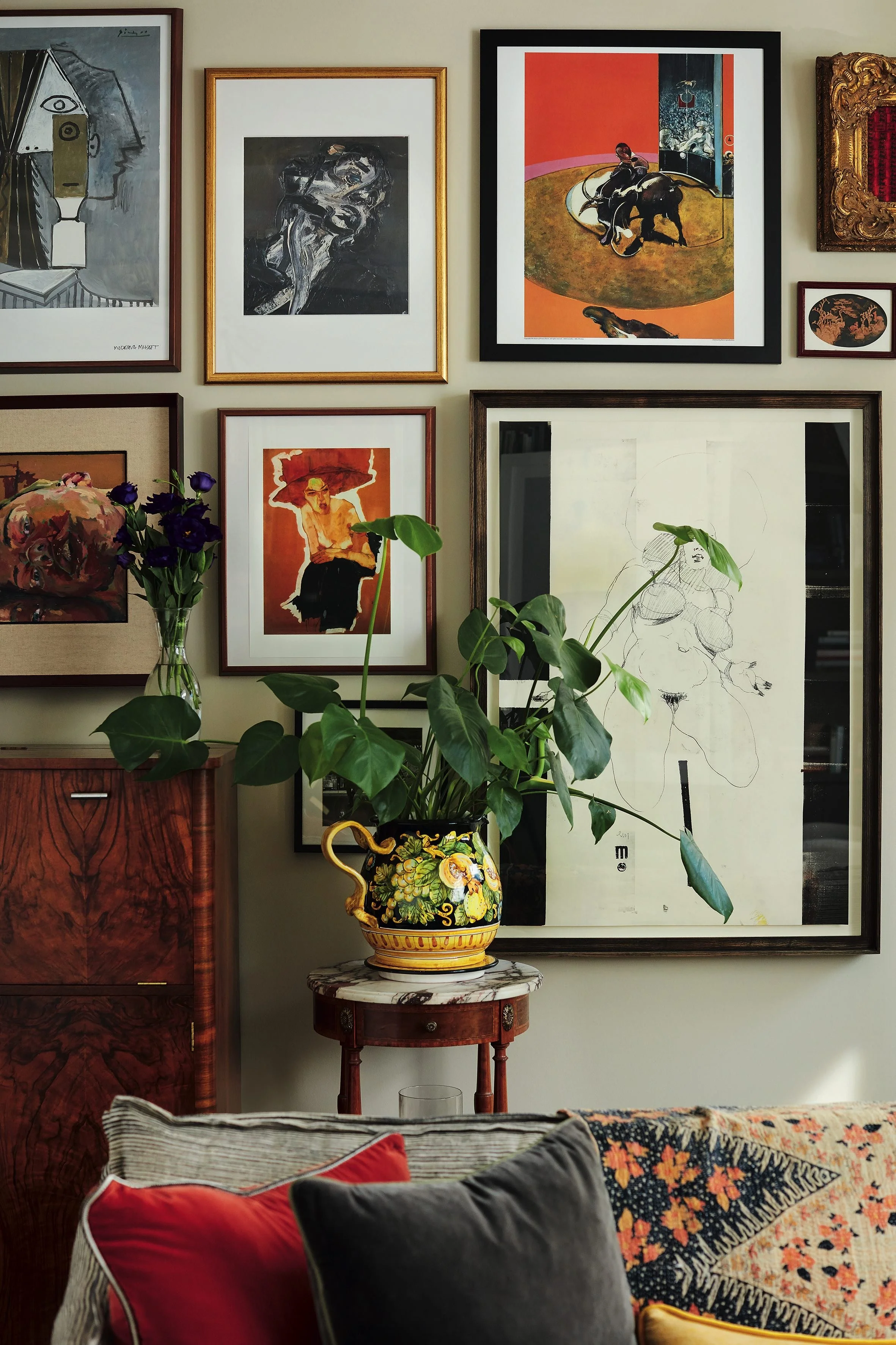 Wall decorated with multiple framed artworks, a china vase with purple flowers on a small wooden table, and a sofa with patterned and solid color pillows.