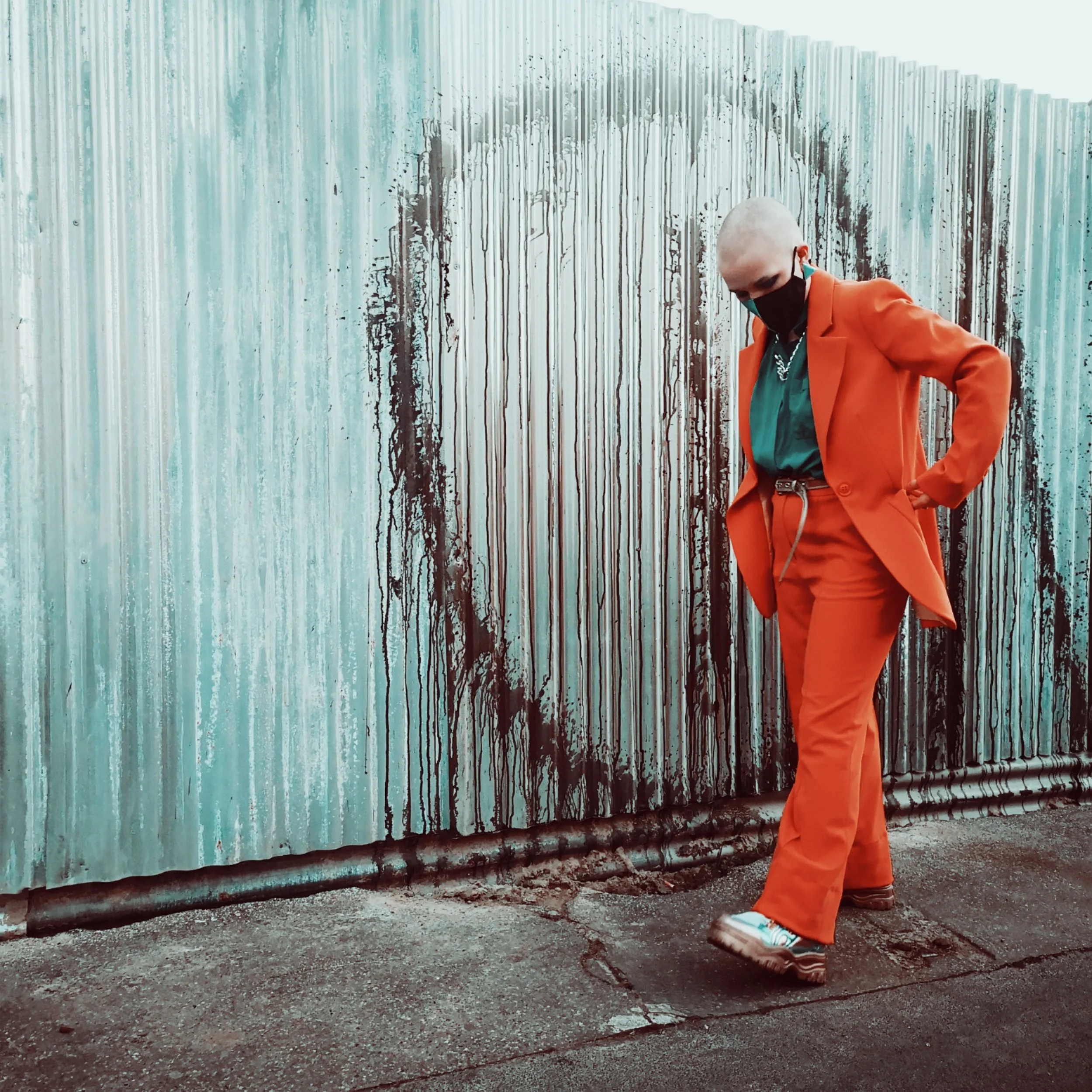 Person with platinum blonde hair wearing a black face mask, orange blazer, teal shirt, orange pants, and striped shoes standing next to a large, weathered, corrugated metal fence with drip patterns.