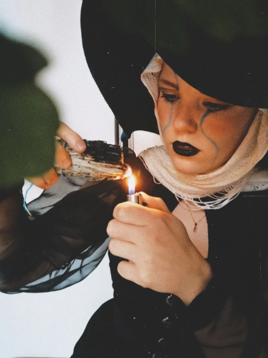 Person dressed as a clown with black makeup and tears painted on their face, wearing a black hood and scarf, is holding a lighter to a piece of paper or fabric with a flame.