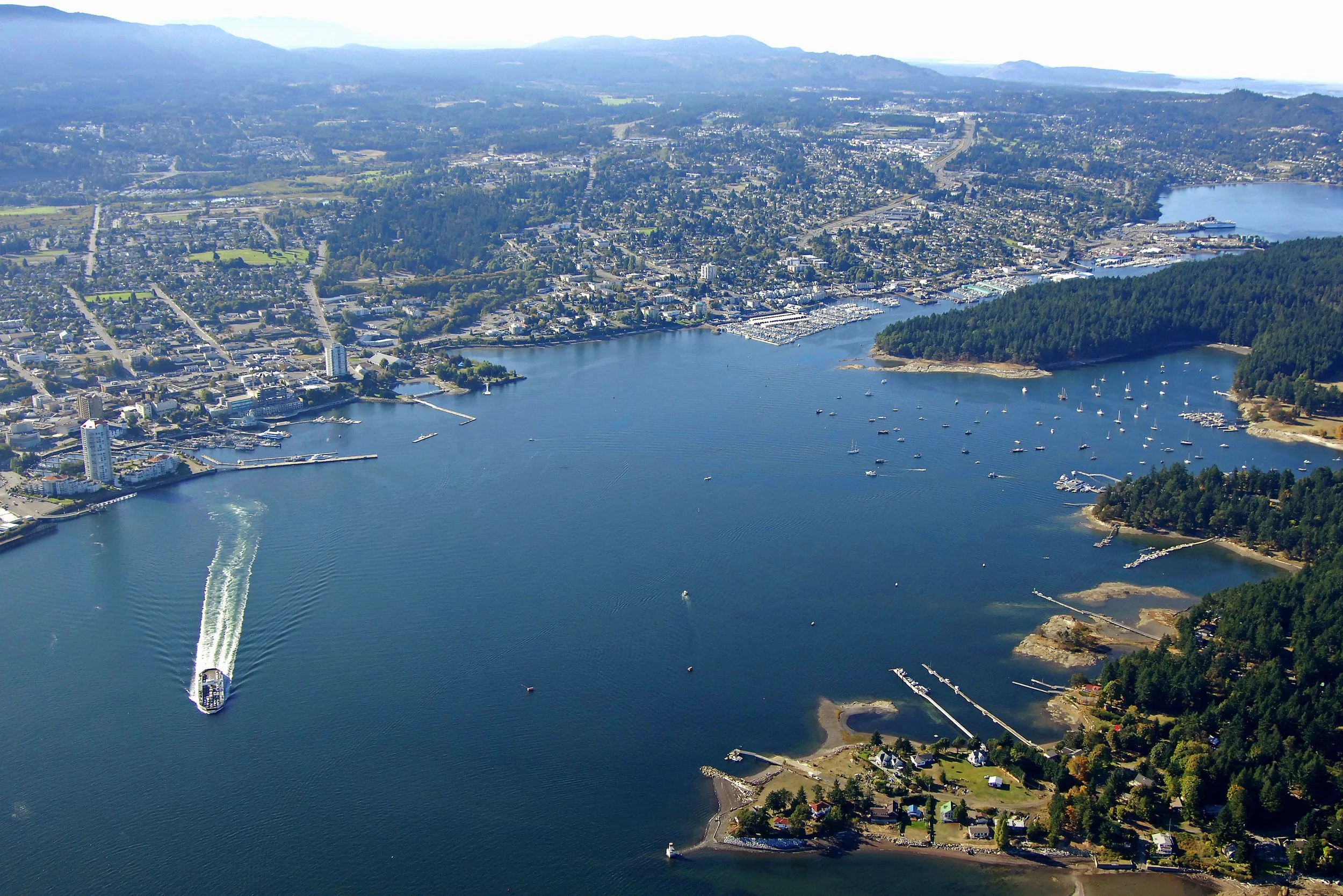 An aerial view of a city by a large body of water with a boat leaving a wake. The city has numerous buildings, roads, and a marina with sailboats, surrounded by forested hills and mountains in the distance.