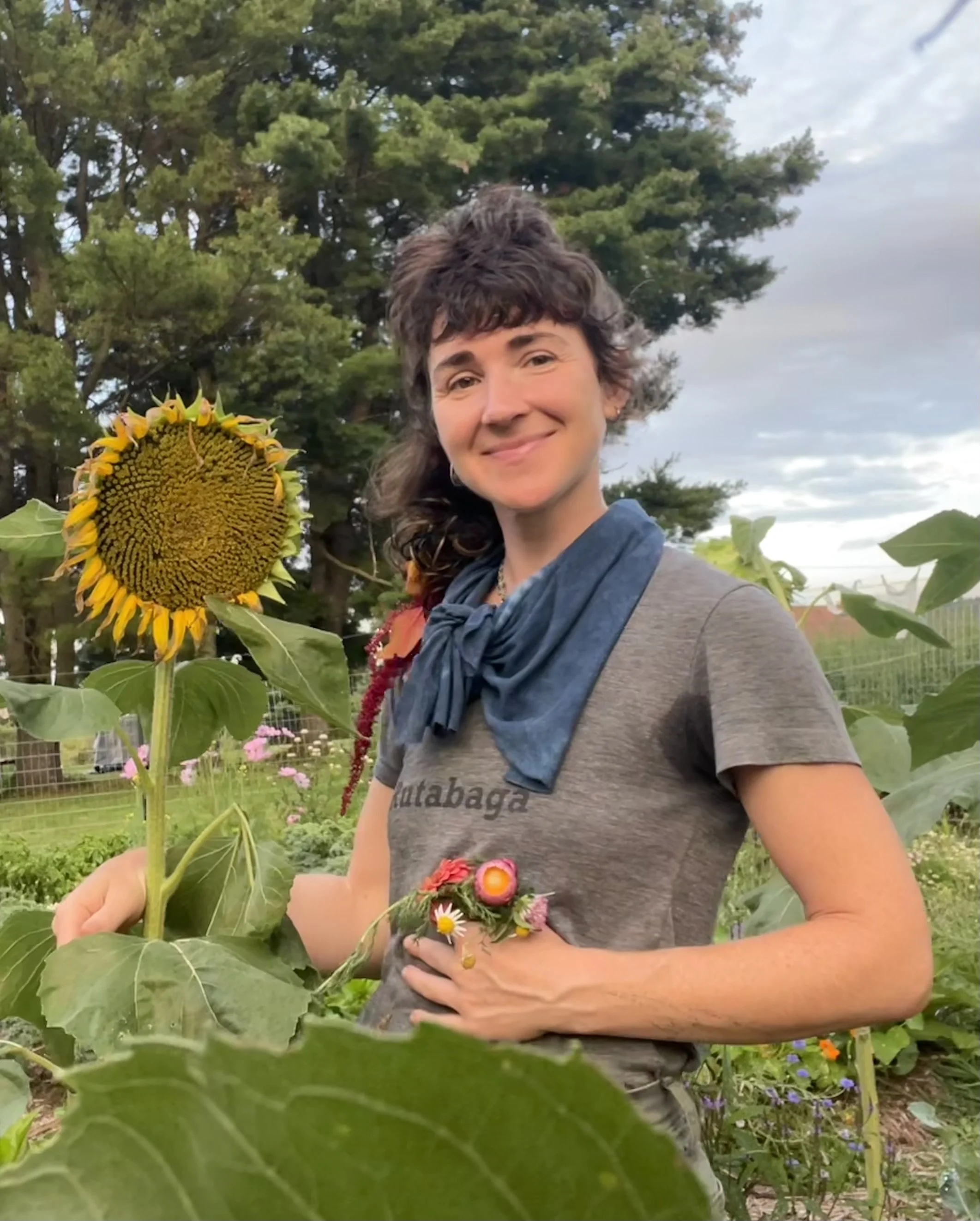 Woman with curly hair smiling in a garden or farm, holding a bouquet of colorful flowers and standing next to a tall sunflower plant.