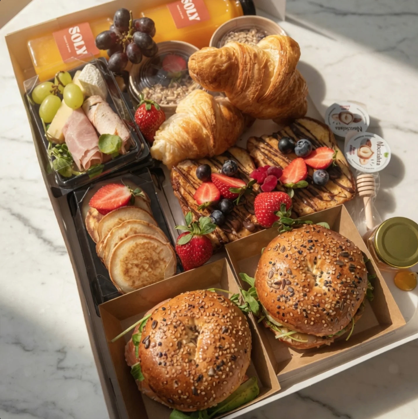 Un plateau de petit-déjeuner avec des viennoiseries, sandwichs, pancakes, fruits frais, jus, fromage, charcuterie, beurre, et confiture