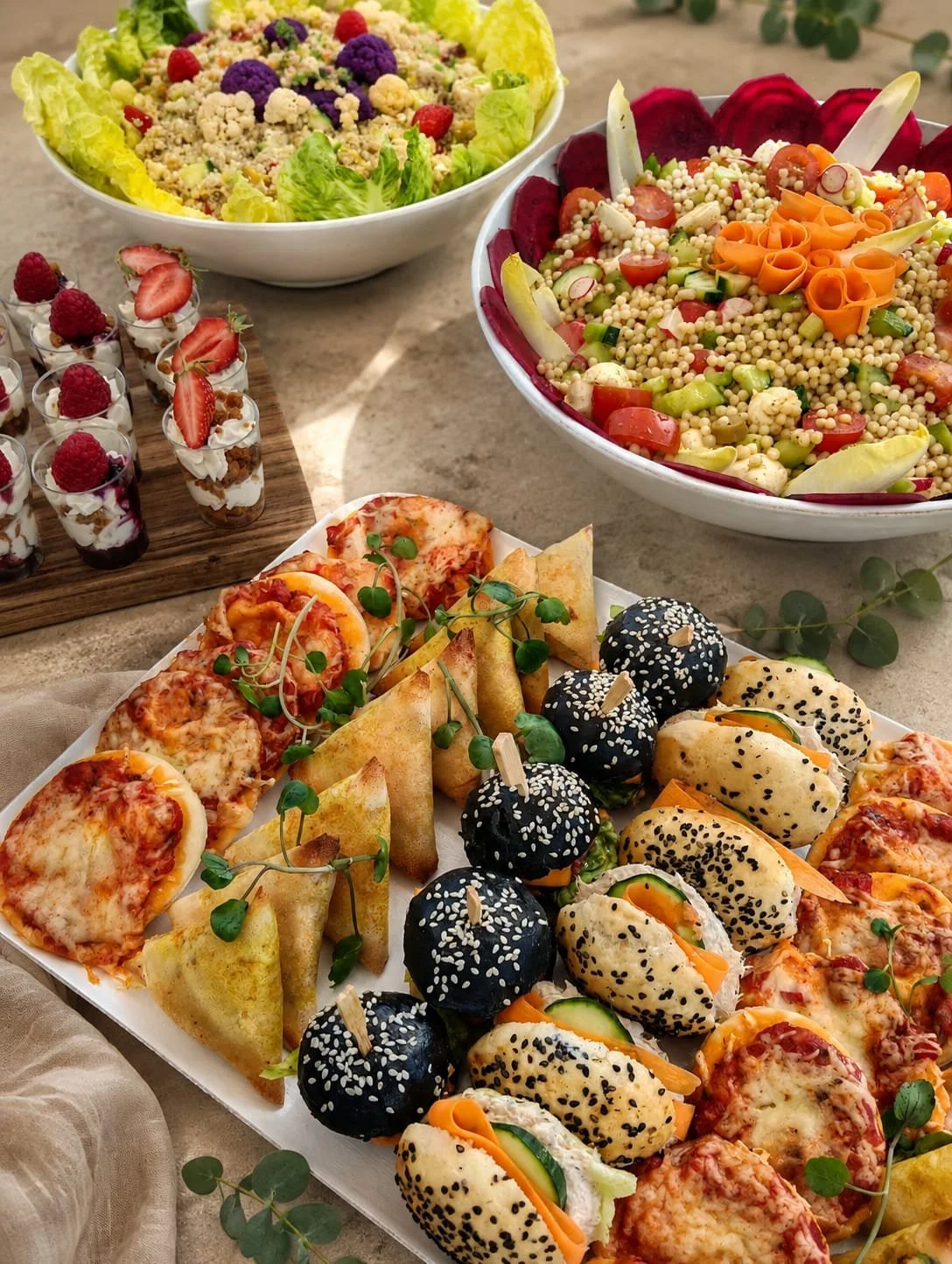 Un buffet de snacks comprenant des pizzas, des mini sandwiches aux graines de sésame, des roulés de légumes, des balls noires avec des wraps, des verrines de dessert aux fruits rouges et des salades de légumes colorés.