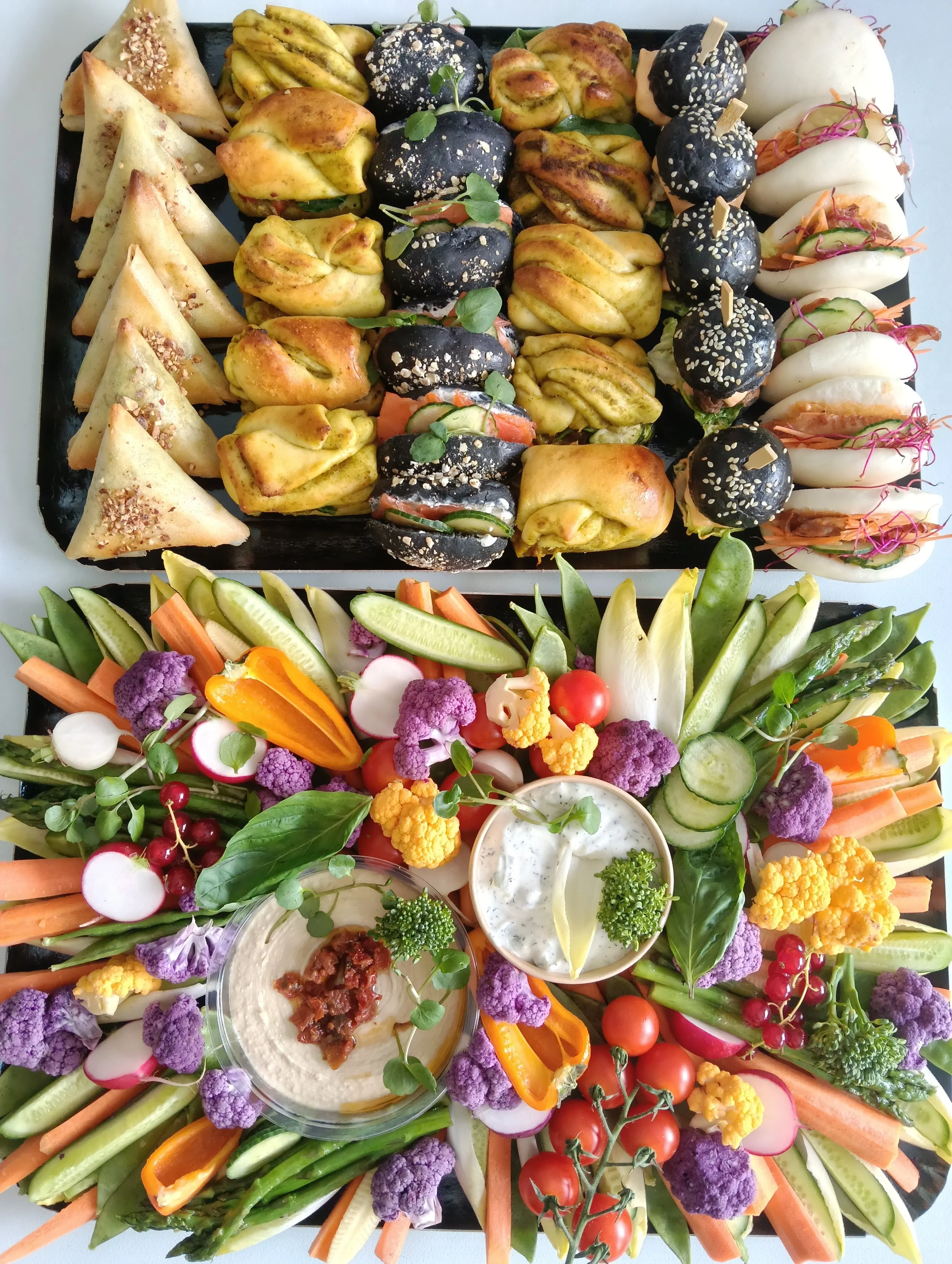 Plateau de légumes et amuse-bouches variés, notamment des légumes crus comme carottes, concombres, radis, tomates cerises, et des apéritifs comme des bouchées de pain farcies, des sphères noires enrobées de graines, des dips et des garnitures végétales.