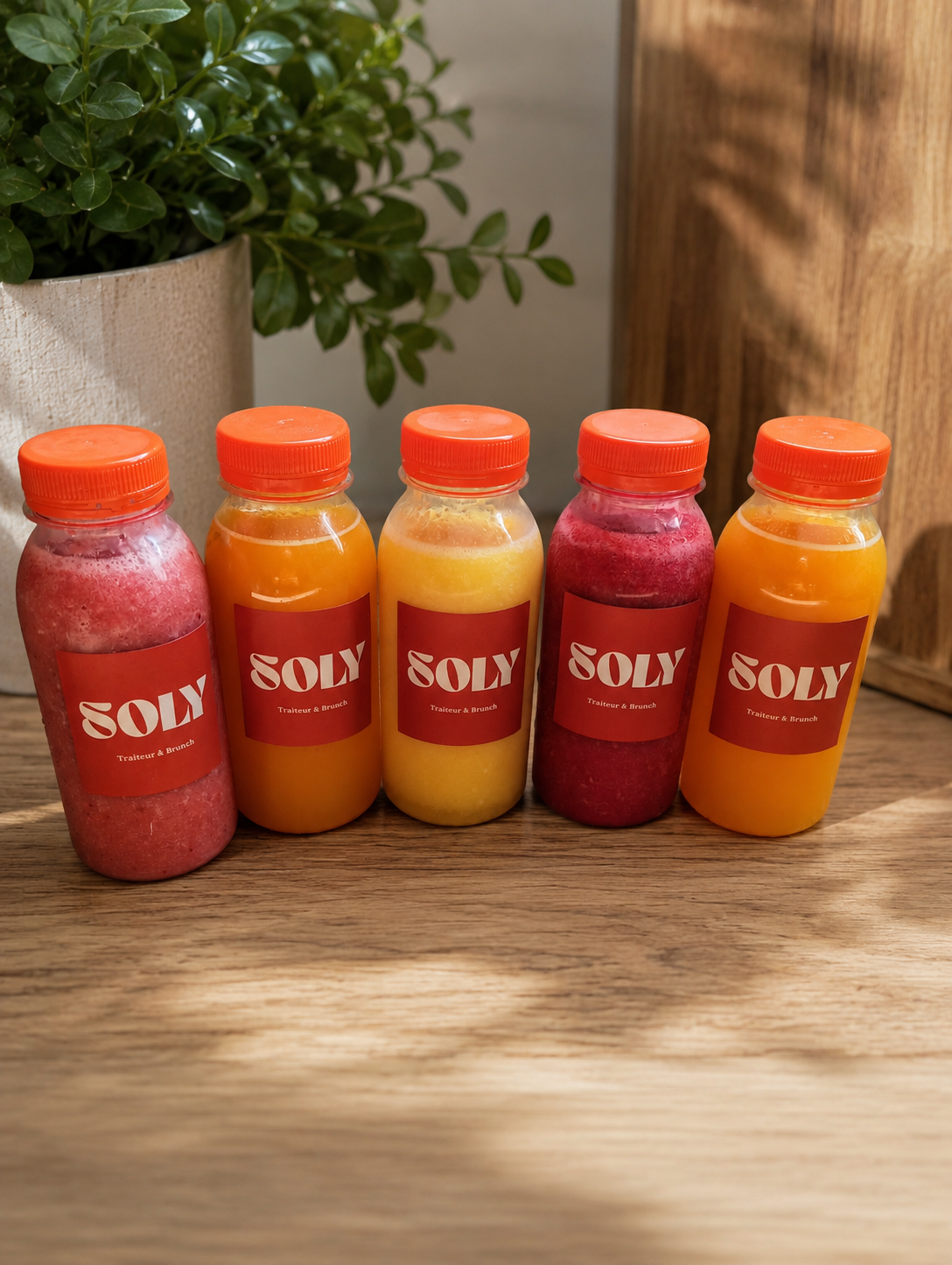 Cinq bouteilles de smoothies aux couleurs variées, avec des étiquettes rouges portant le nom 'SOLY' et la mention 'Traiteur & Brunch', posées sur une table en bois, avec un pot de plantes vertes en arrière-plan.