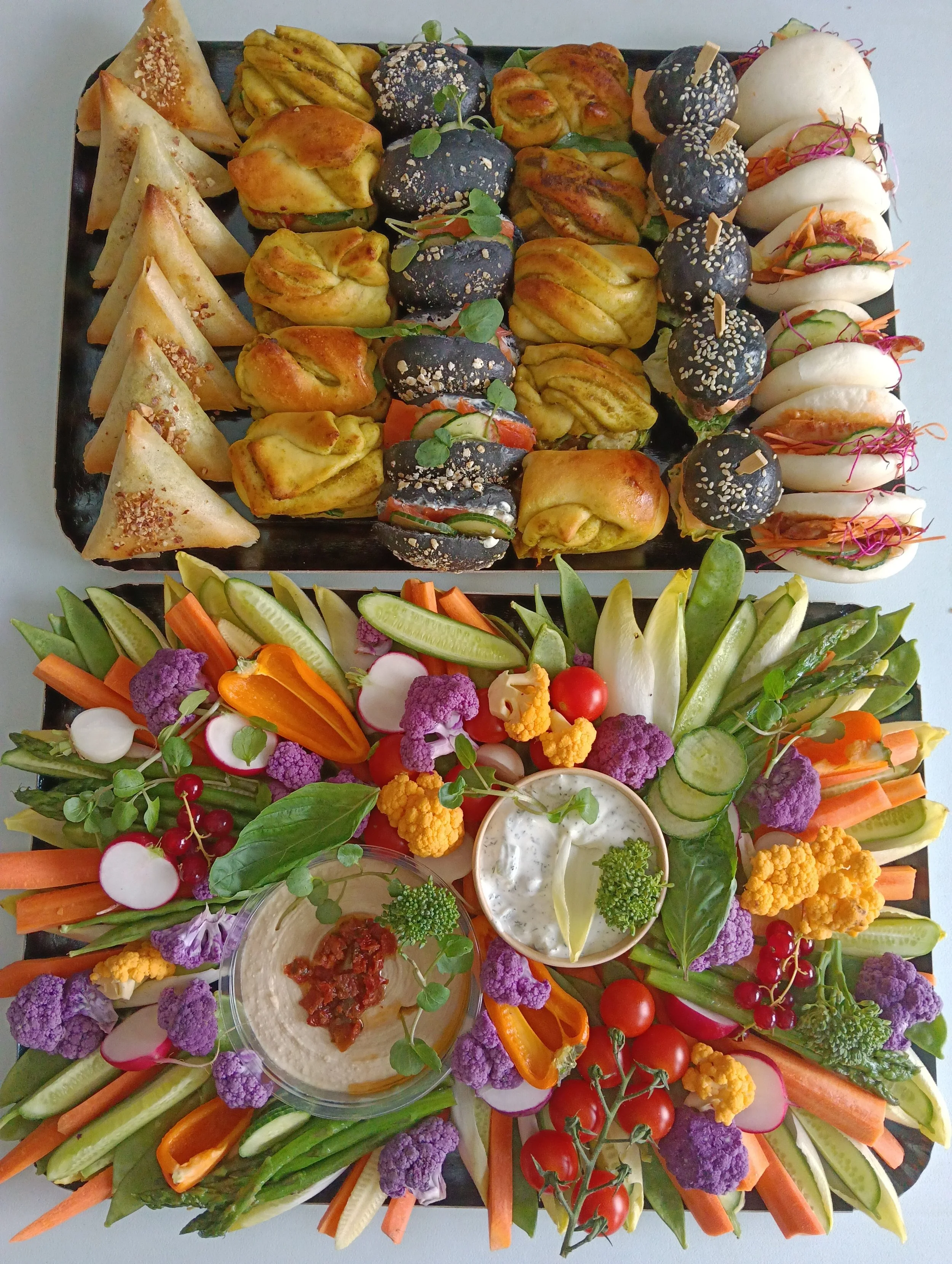 Plateau avec une variété de appetizers et légumes frais, comprenant des sushis, des boulettes noires garnies de graines de sésame, des samoussas, des dips, et des légumes crus coupés en julienne, décoré de microgreens et fleurs violettes.
