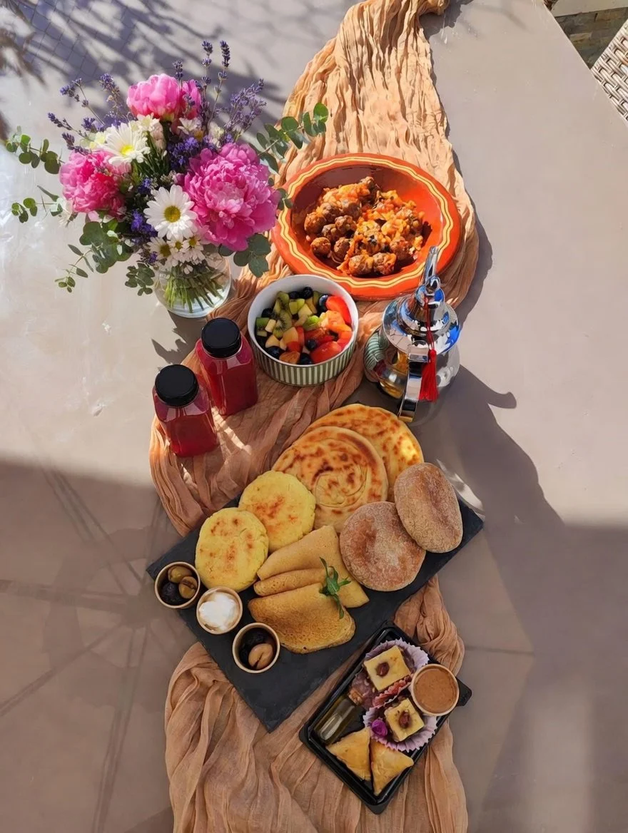 Table dressée avec un bouquet de fleurs, un plat de viande en sauce, un bol de fruits coupés, plusieurs types de pains et pâtisseries, et deux bouteilles de jus de fruit.
