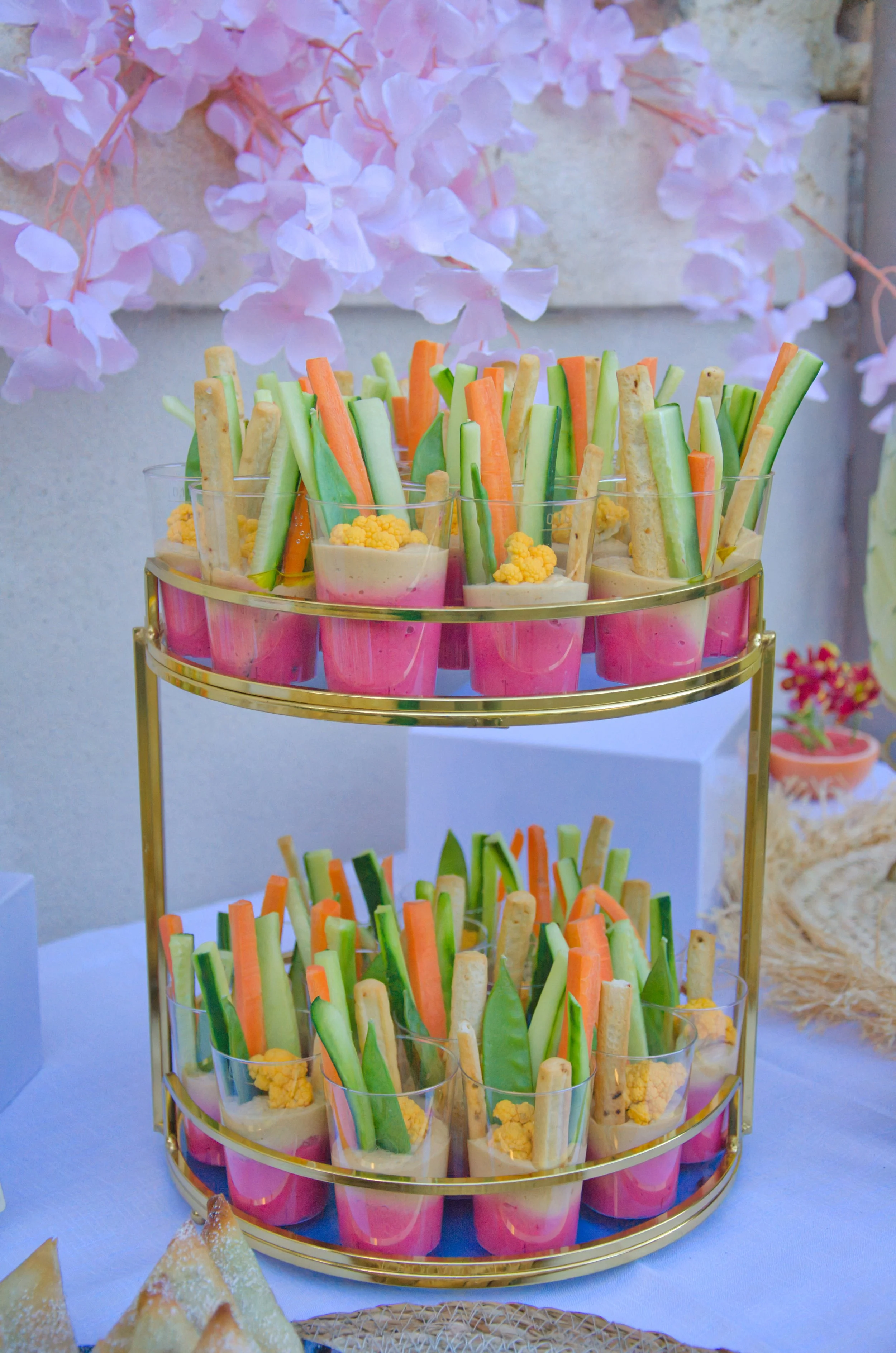 Verres transparents contenant des bâtonnets de légumes verts, oranges et beige, garnis de petits morceaux de chou-fleur jaune, disposés sur un support à plusieurs niveaux, avec des décorations florales roses en arrière-plan.
