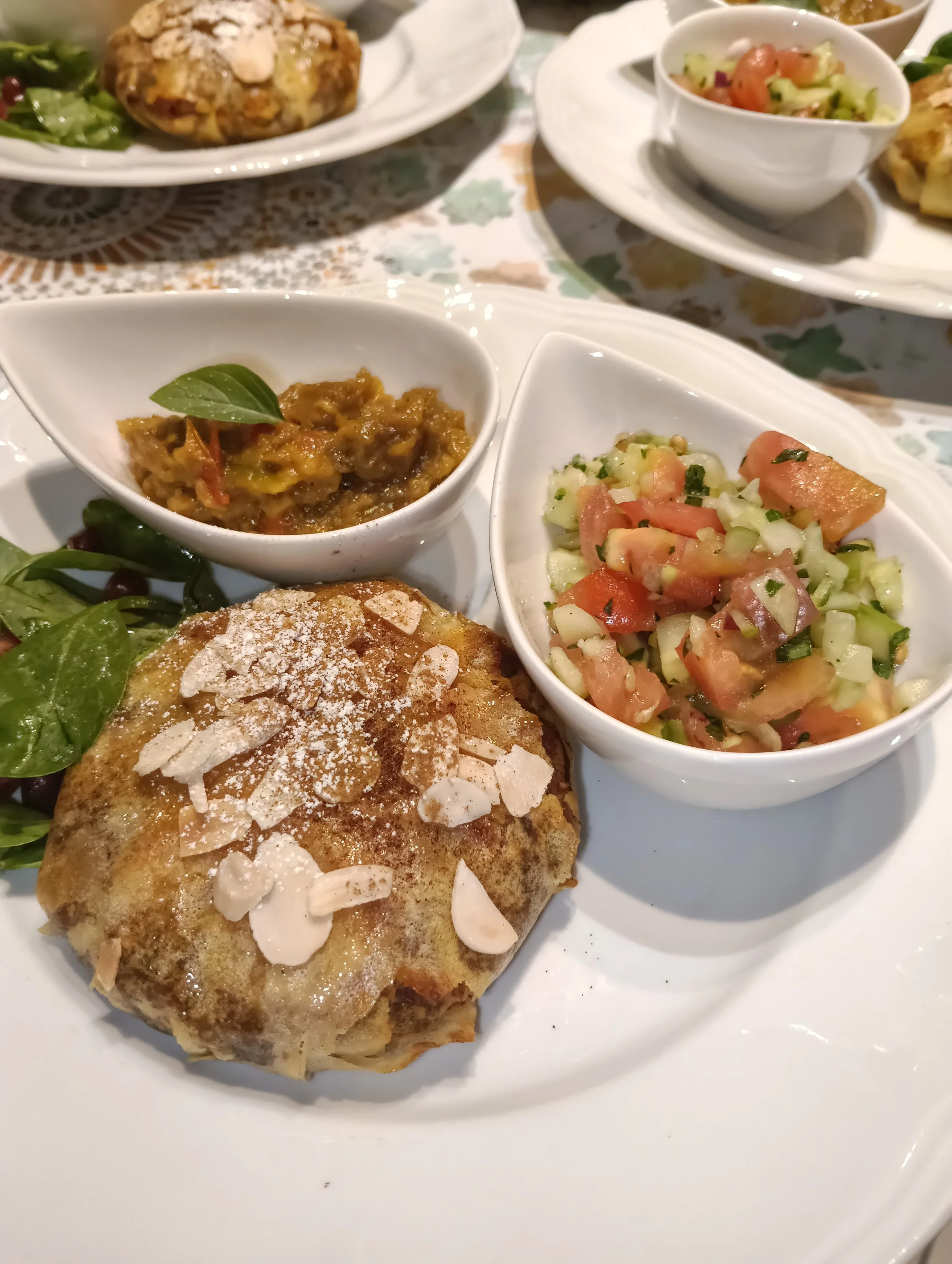 Assiette avec un morceau de pain ou galette garni d'amandes effilées, accompagné d'une petite salade verte. En arrière-plan, plusieurs petits bols avec des salades et autres plats.
