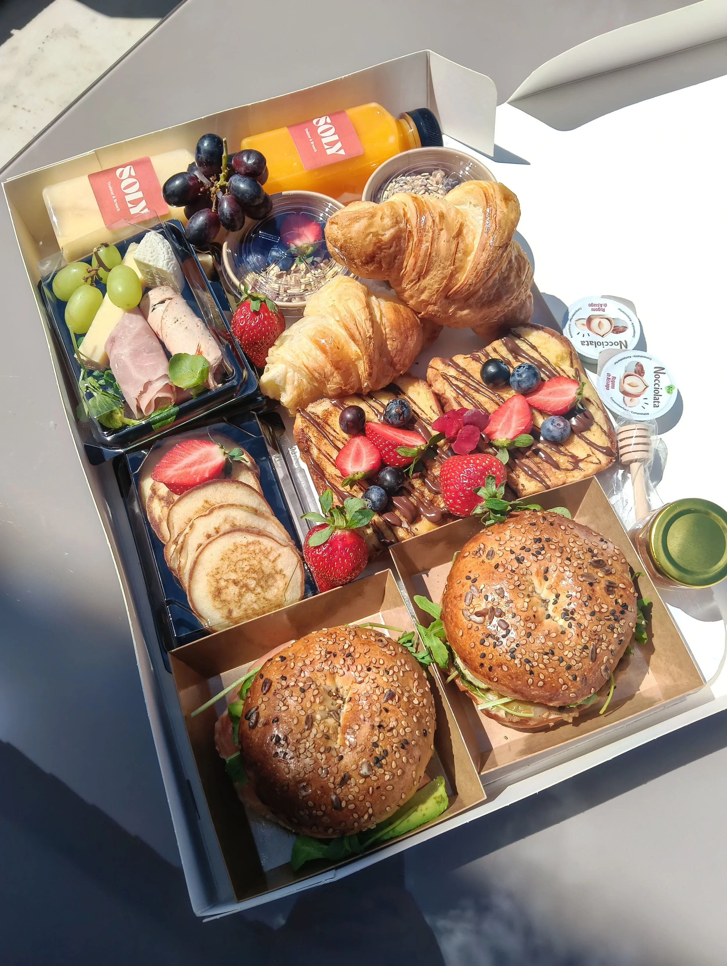 Un plateau de petit-déjeuner avec des croissants, des fruits frais comme des fraises, des raisins, un sandwich, des bouteilles de jus, des yaourts et des chips de noix de coco.