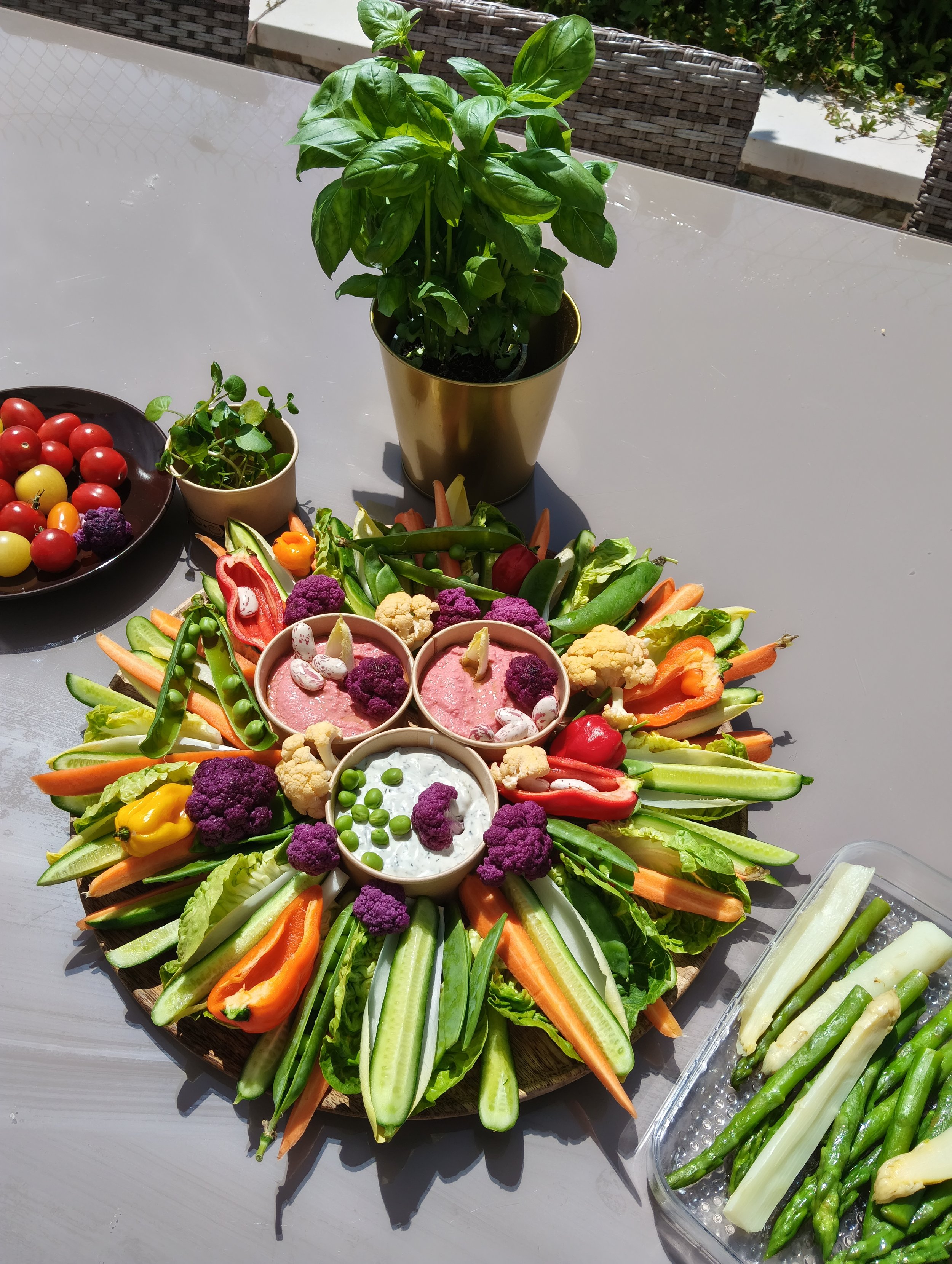 Un plateau de légumes coupés en bâtonnets, comprenant concombre, carotte, poivron, et chou-fleur, entouré de petites coupes avec des dips et un pot de basilic.